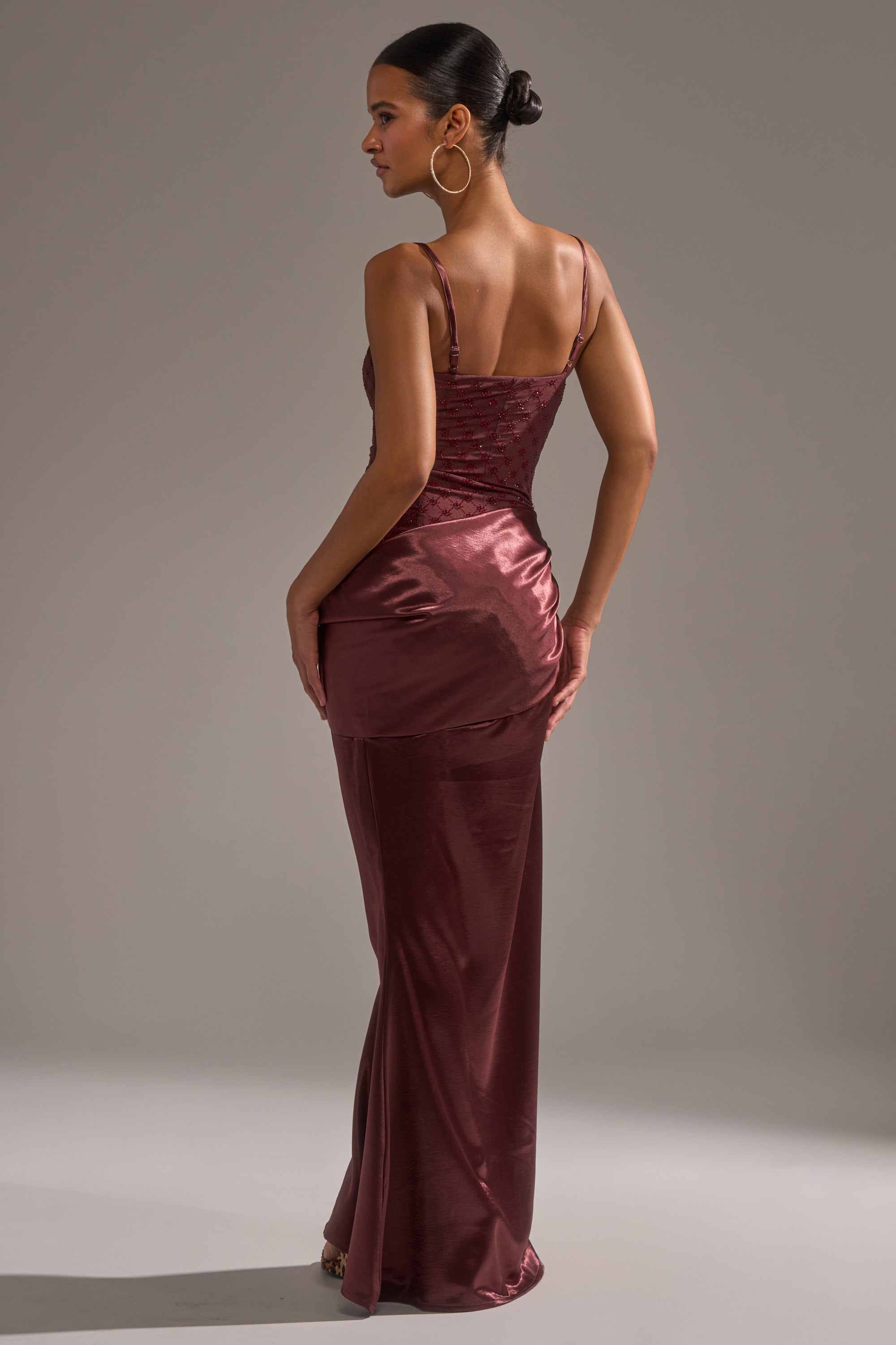 A woman faces away, wearing the RUNAWAY SATIN MESH MAXI DRESS with spaghetti straps. Her hair is in a low bun, accented by large hoop earrings. The background is plain and neutral.