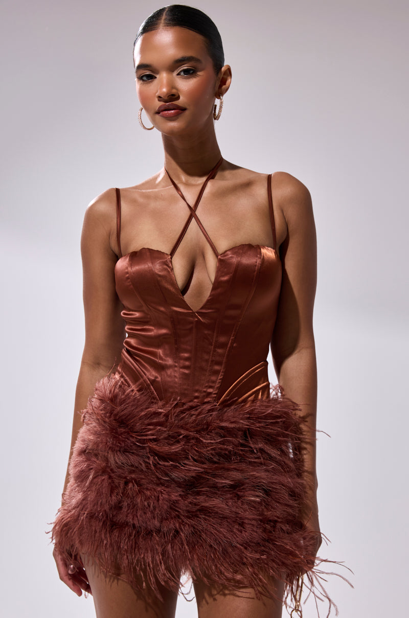 A woman poses against a plain background wearing the SOMETHIN EXTRA FEATHER MINI DRESS—a shiny, rust mini dress with a deep V-neck, spaghetti straps, and feathered skirt—styled with hoop earrings and a sleek hairstyle for a chic look.