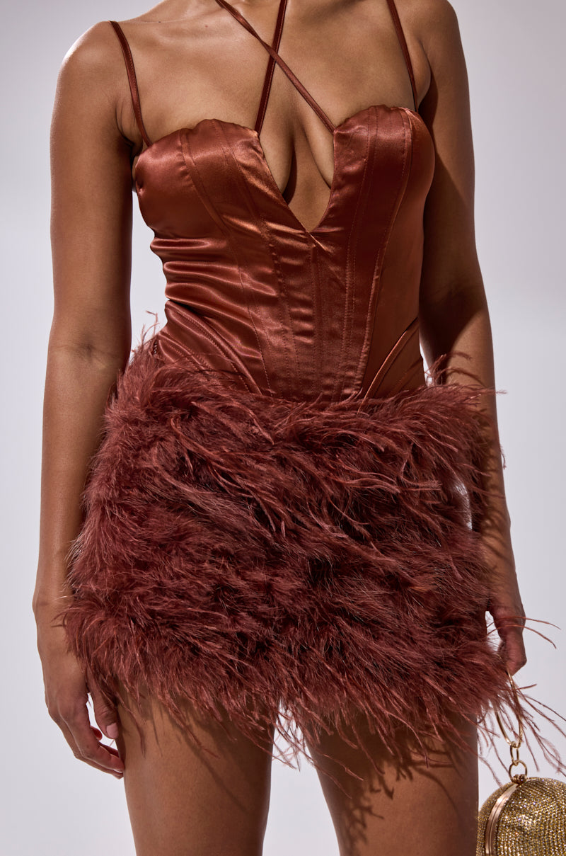 A woman wears the SOMETHIN EXTRA FEATHER MINI DRESS, a fitted deep rust satin mini with a plunging neckline, crisscross straps, and matching feather accents, paired with a small gold handbag.