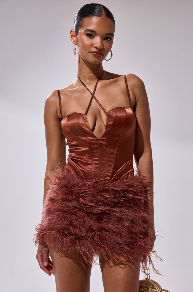 A woman wears the SOMETHIN EXTRA FEATHER MINI DRESS, a shiny rust mini with a deep neckline, spaghetti straps, and playful feathered skirt. With slicked-back hair and hoop earrings, she’s ready for any cocktail party.
