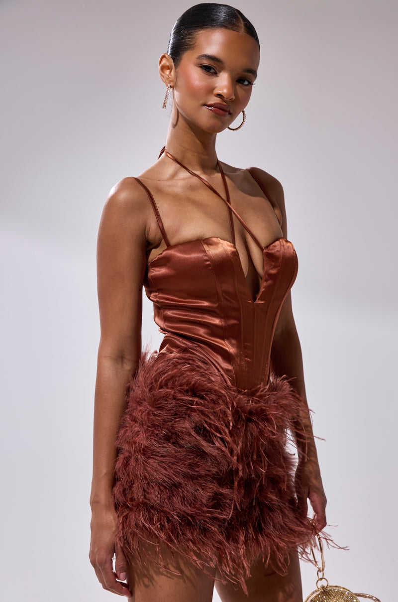 A woman stands confidently in the SOMETHIN EXTRA FEATHER MINI DRESS, featuring satin fabric, spaghetti straps, and a feathered skirt detail. She styles her hair back and accessorizes with hoop earrings against a plain background.