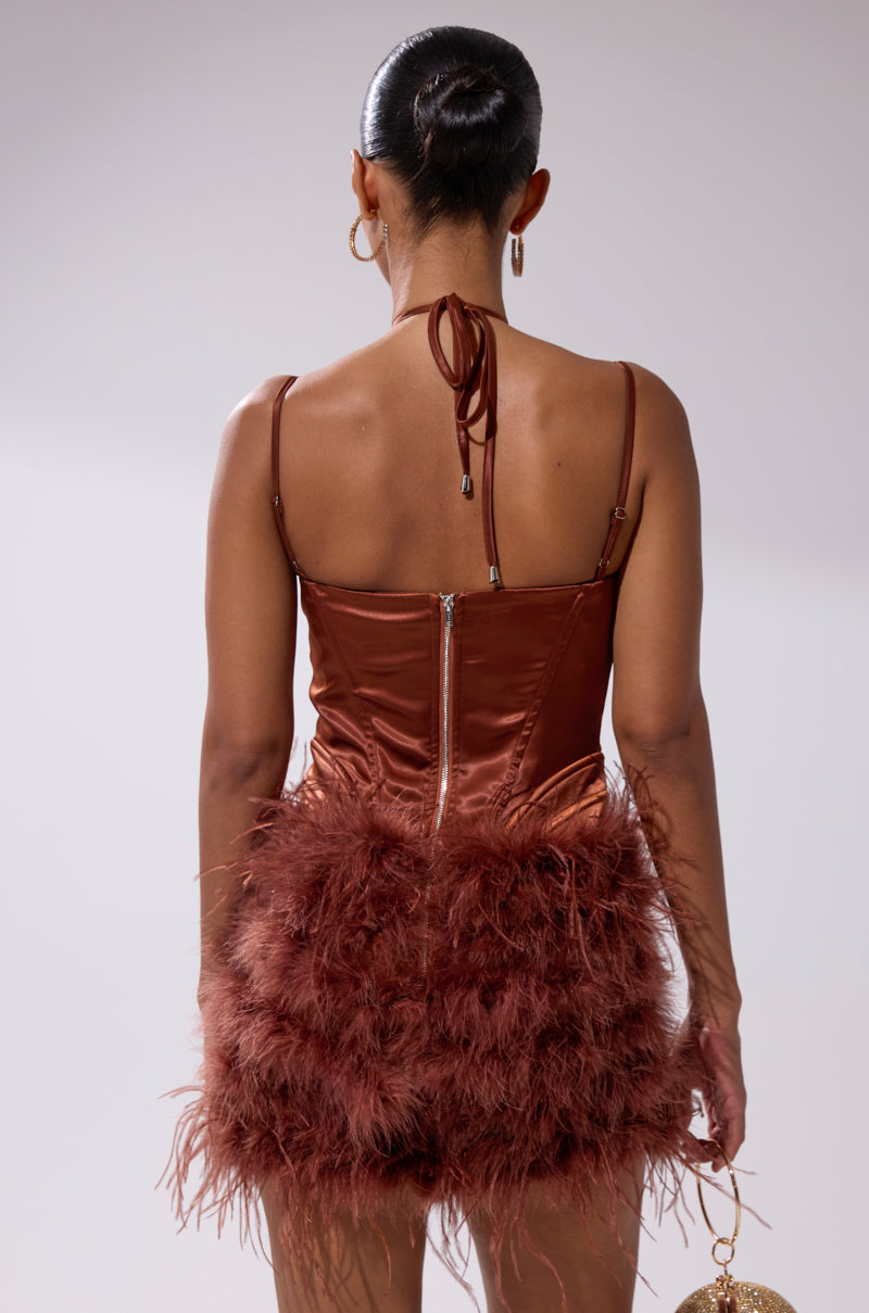 A woman with a sleek bun models the SOMETHIN EXTRA FEATHER MINI DRESS, a brown satin mini with feathered trim, thin straps, and a subtle corset bodice. Seen from the back, she holds a small round purse against a neutral background.