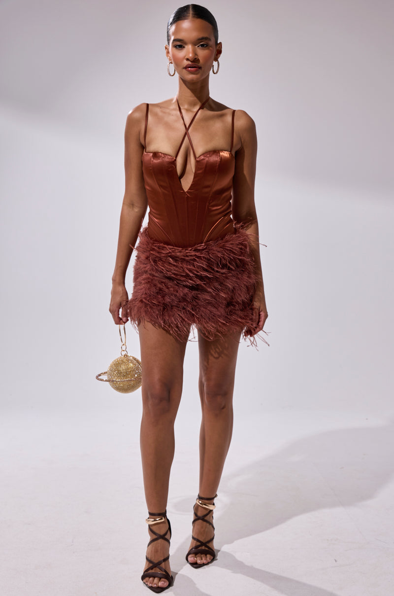 A woman wearing the SOMETHIN EXTRA FEATHER MINI DRESS in rust satin with a halter neckline and feathered skirt, paired with strappy heels, hoop earrings, and a small round gold purse stands against a plain white background.