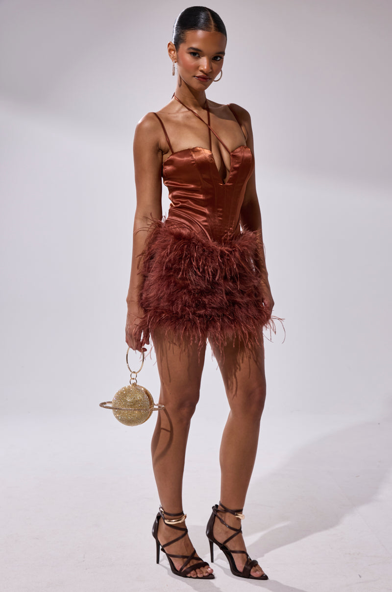 A woman stands confidently in the SOMETHIN EXTRA FEATHER MINI DRESS, featuring a copper hue and satin corset bodice, paired with strappy black heels and a round gold handbag. Her hair is slicked back and she wears dark lipstick against a plain background.
