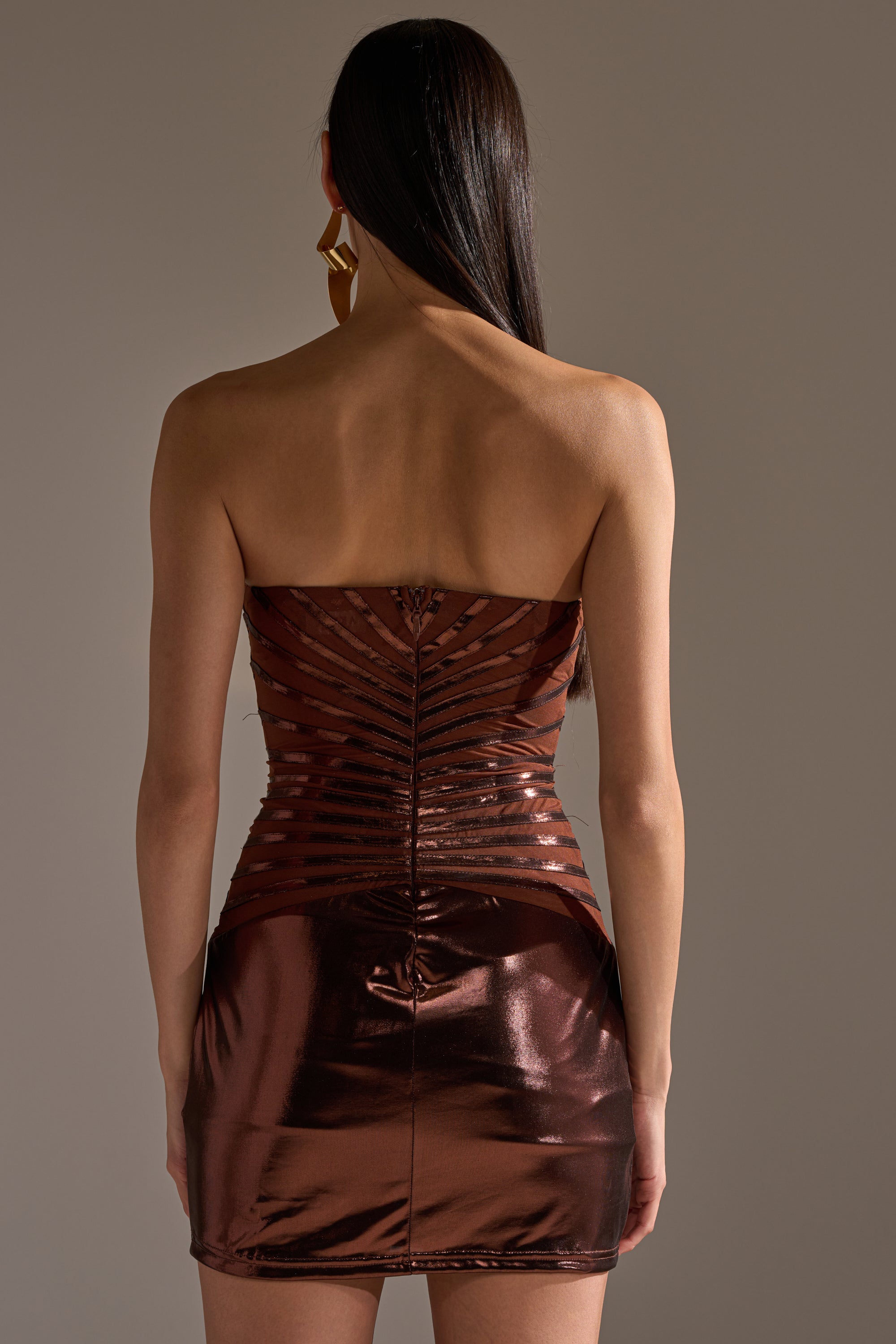 A woman with long dark hair, seen from behind, wears the ON THE MOVE BRONZE MINI DRESS, a strapless mini with ruched and sheer striped details. She accessorizes with a large geometric gold earring in one ear. 4