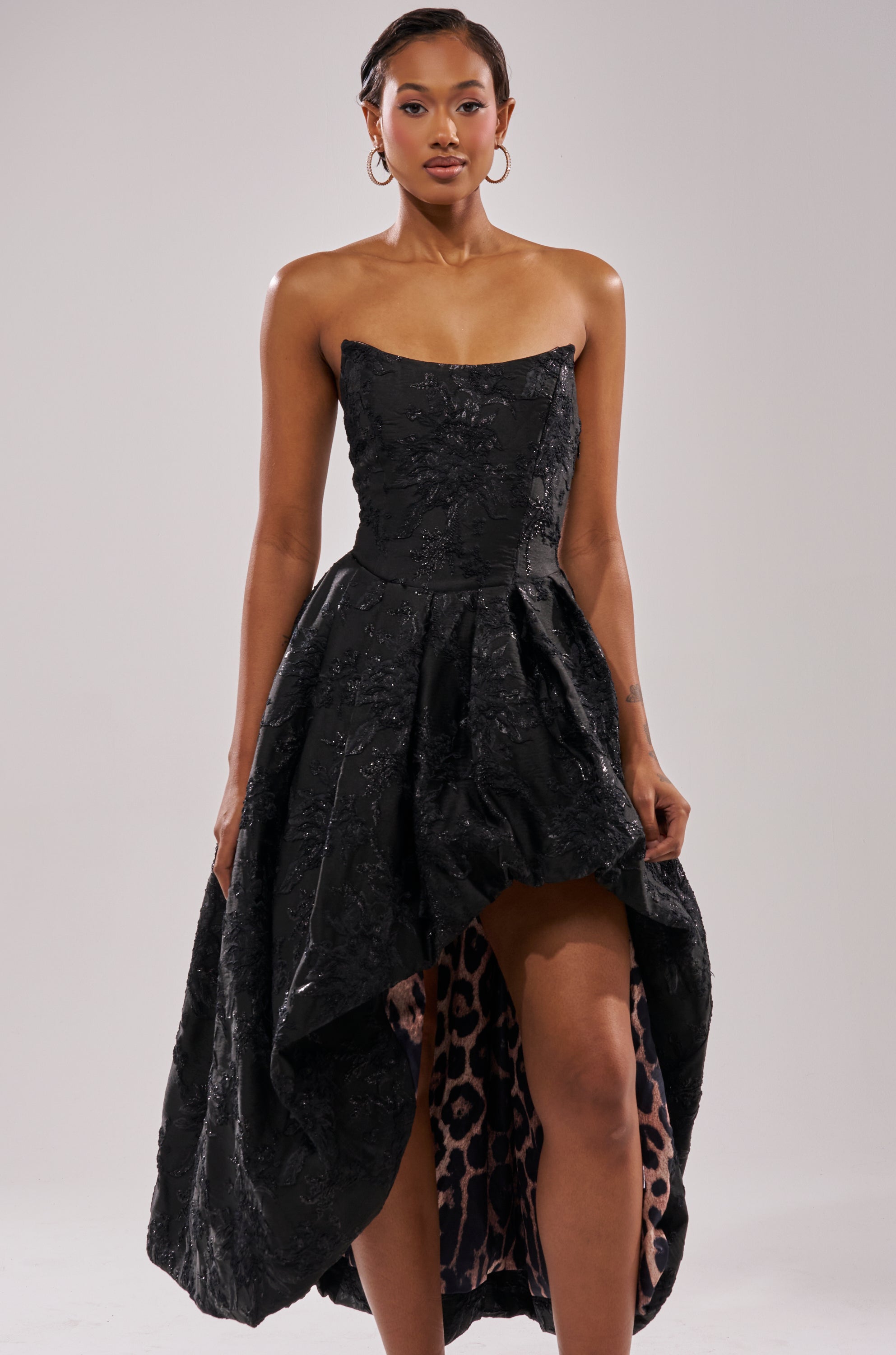 A woman models the WILD THANG PRINTED HI LOW DRESS, a strapless jacquard dress with subtle florals and visible leopard-print lining, standing against a plain light background. 1