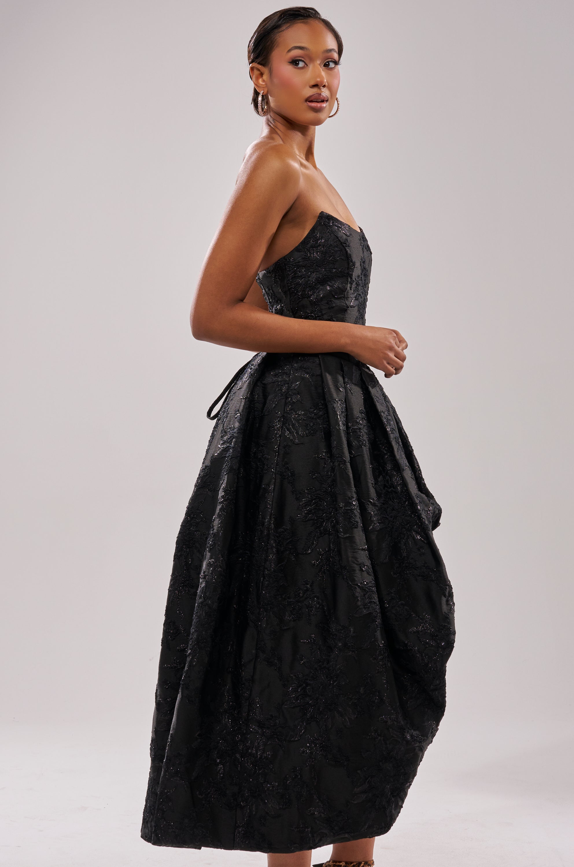 A woman poses sideways in the WILD THANG PRINTED HI LOW DRESS, featuring a strapless design and textured black full skirt, hands clasped, gazing toward the camera against a plain light background. 2