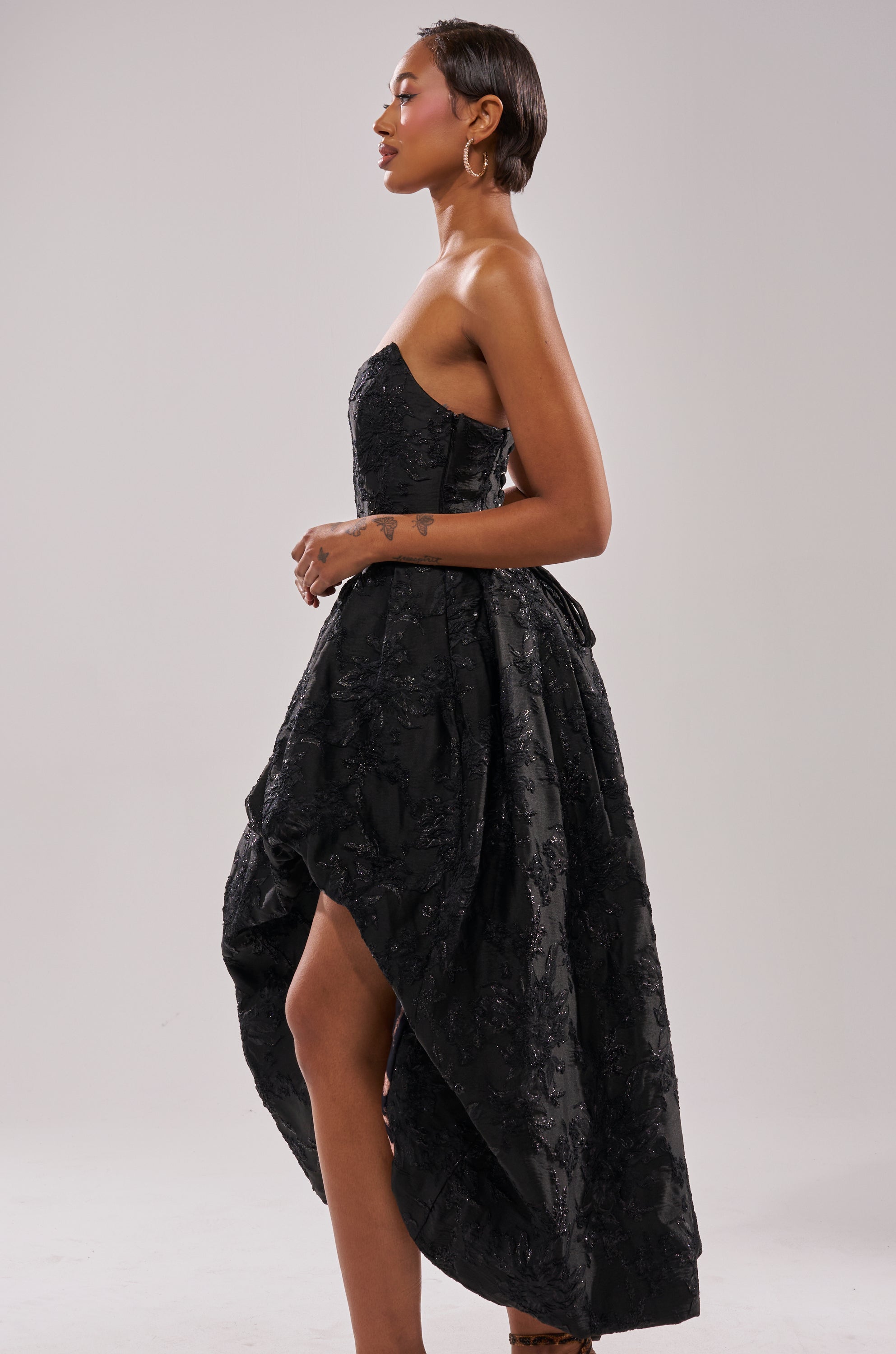 A woman with short hair and hoop earrings stands in profile against a light gray background, confidently wearing the WILD THANG PRINTED HI LOW DRESS, which features a strapless design and textured black hi low hem. 3