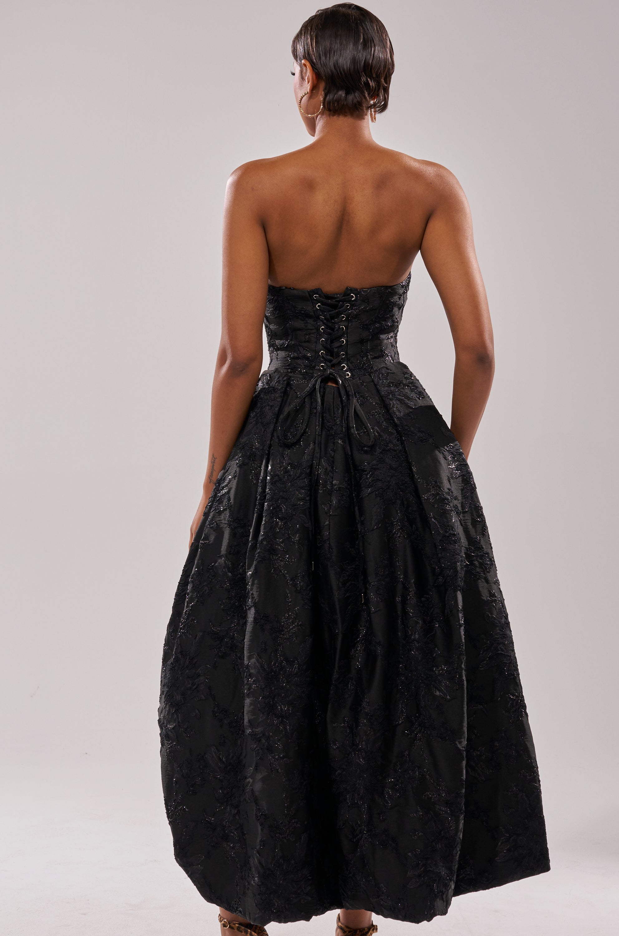 A woman with short, close-cropped hair faces away, wearing the WILD THANG PRINTED HI LOW DRESS featuring a strapless design, lace-up back detailing, and a textured, voluminous skirt against a plain light background. 4