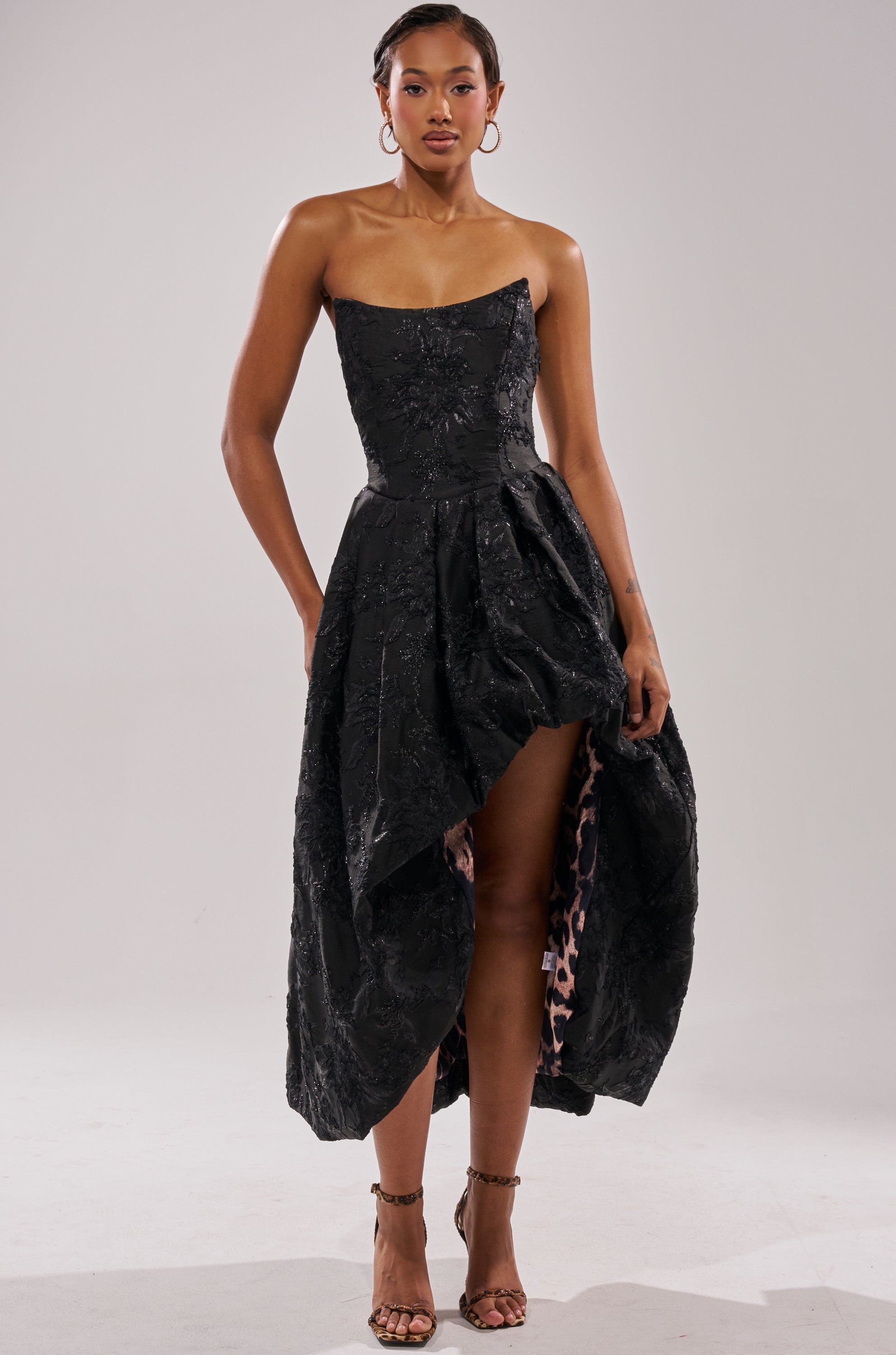 A woman poses confidently in strappy heels, wearing the WILD THANG PRINTED HI LOW DRESS—an elegant strapless black dress featuring embossed jacquard texture and a leopard print lining, set against a plain background. 5
