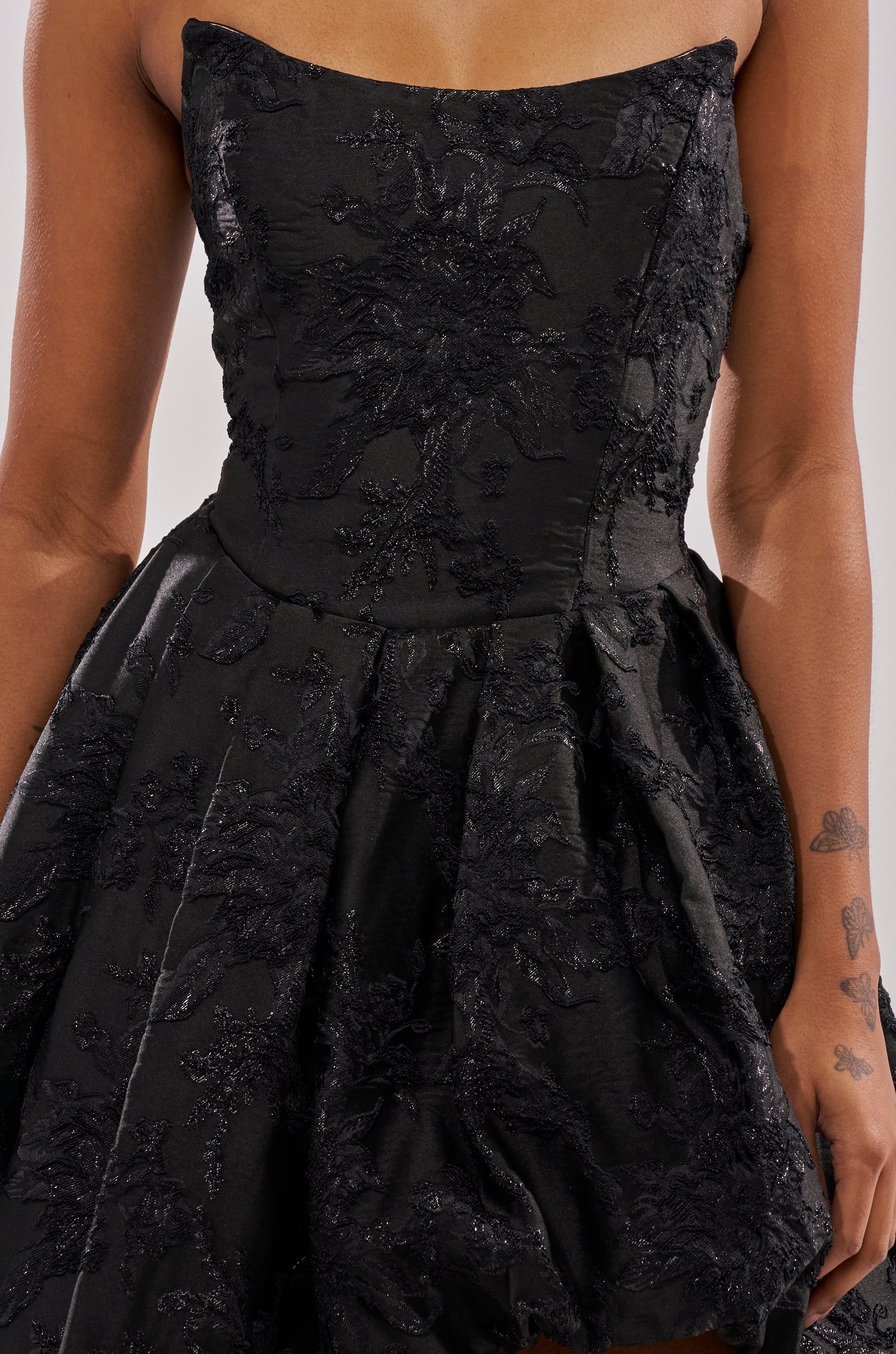 A person wears the WILD THANG PRINTED HI LOW DRESS, a strapless black evening dress with an embossed jacquard finish and flared skirt; their arms are bare, featuring visible floral tattoos on one arm. 6