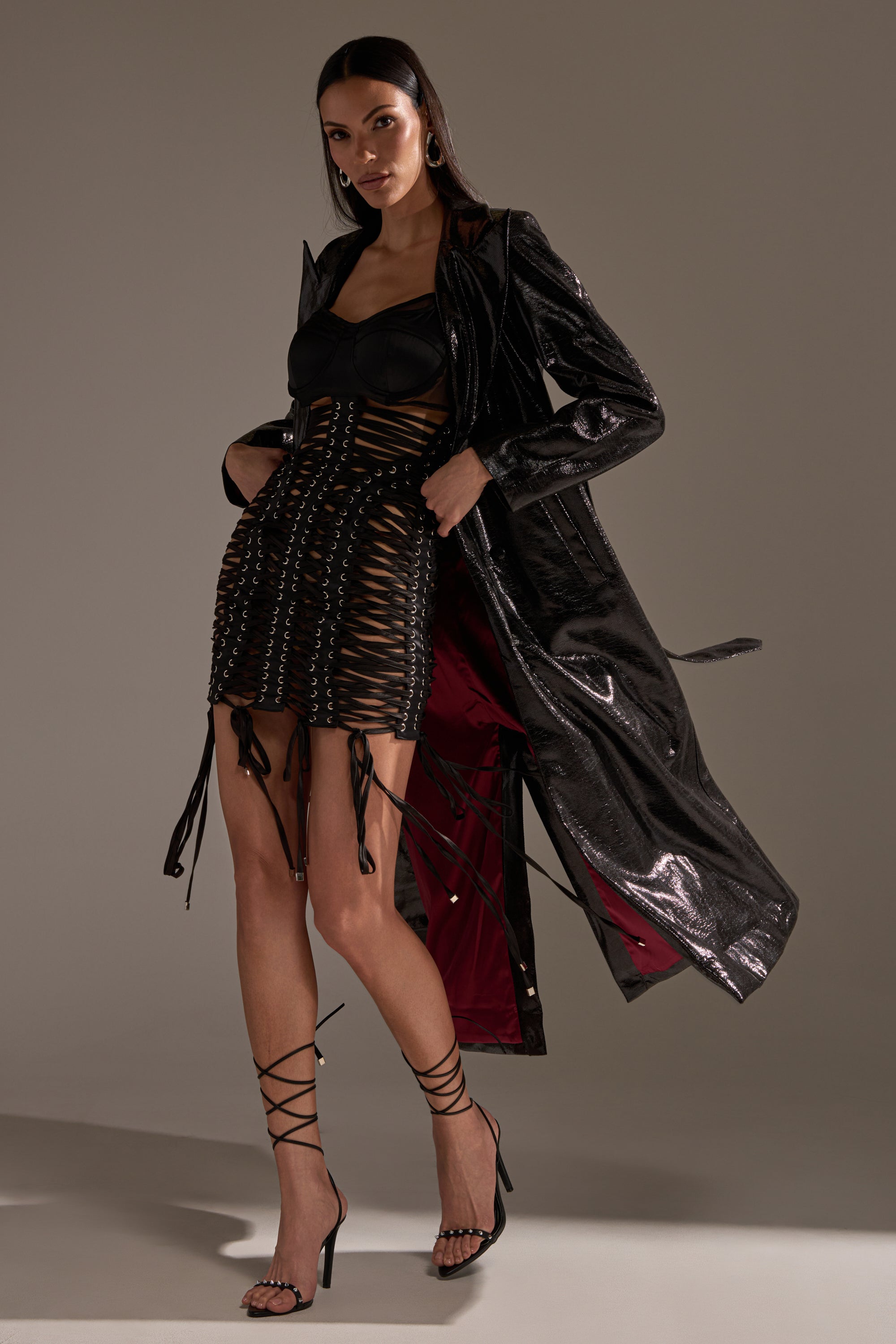 A woman poses in the UNBREAKABLE SATIN LACE UP MINI DRESS, paired with a black shiny trench coat and strappy high-heeled sandals. Her straight, loose hair frames her face as she stands against a plain, neutral background.
