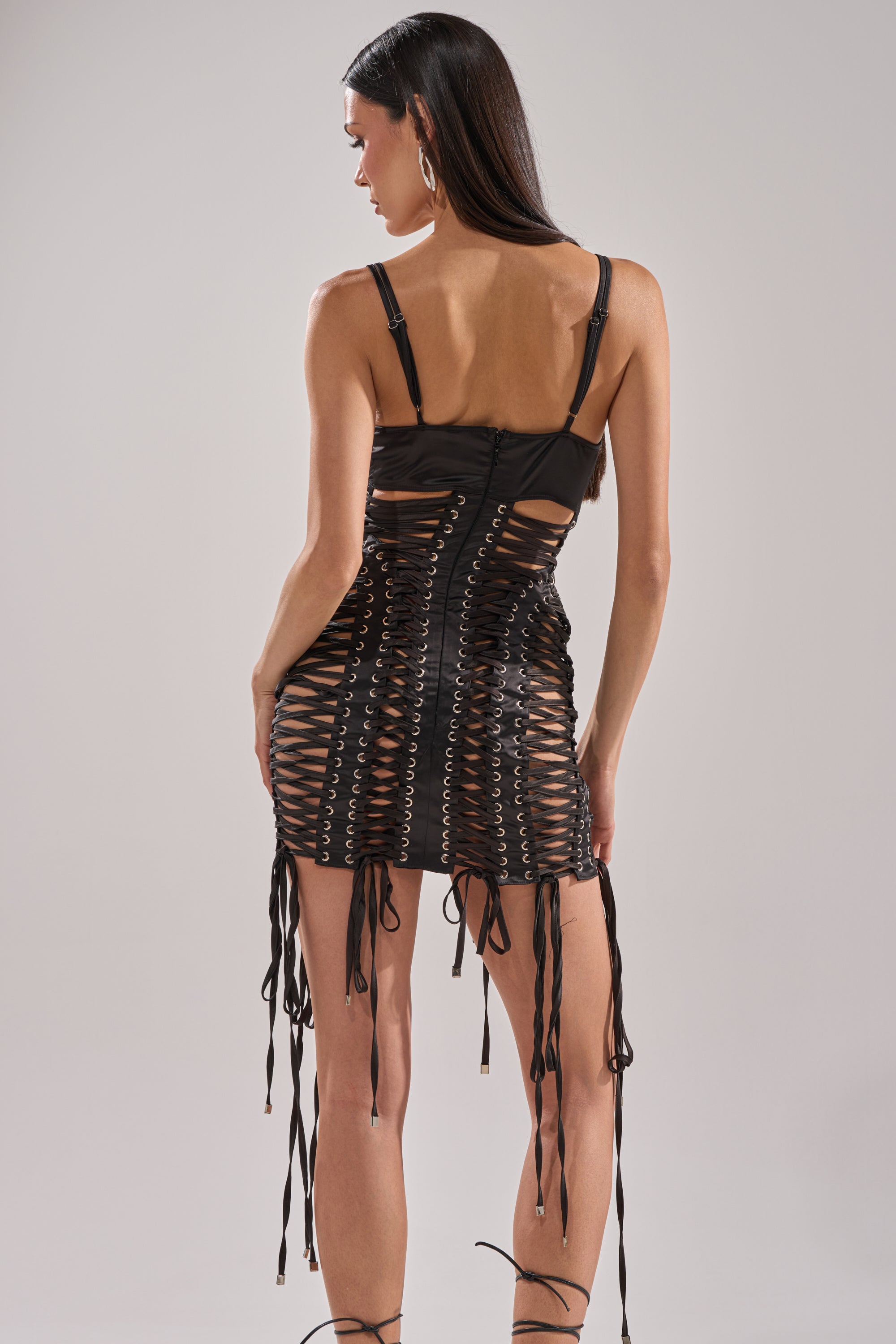 A woman with long dark hair models the UNBREAKABLE SATIN LACE UP MINI DRESS, a fitted black stretch satin piece with intricate lace-up detailing and dangling strings on the back, posed against a plain light background.