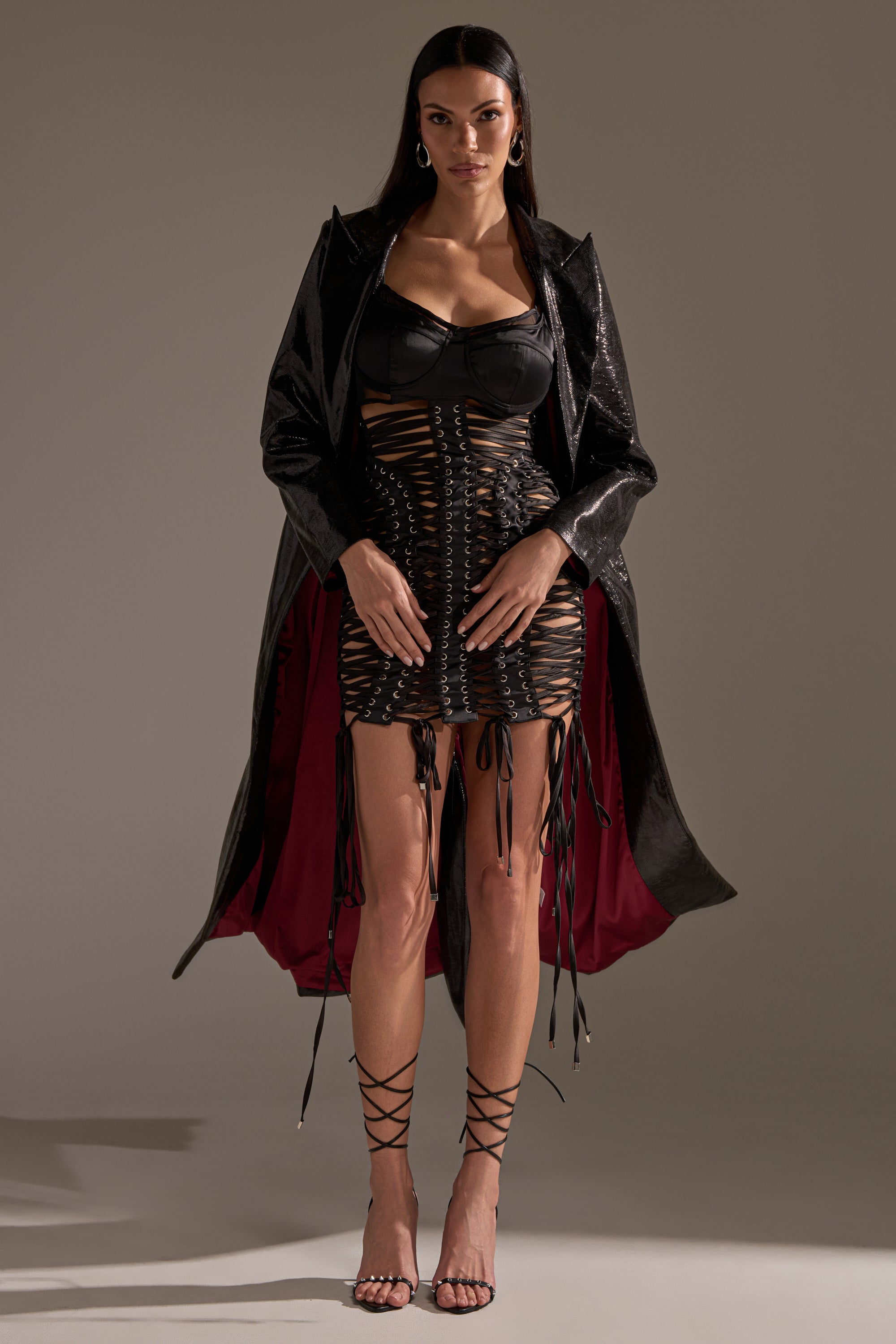 A woman wears the UNBREAKABLE SATIN LACE UP MINI DRESS with a black bra top, long black coat with red lining, strappy black heels, and large hoop earrings. Her long dark hair and bold accessories complete the striking look.