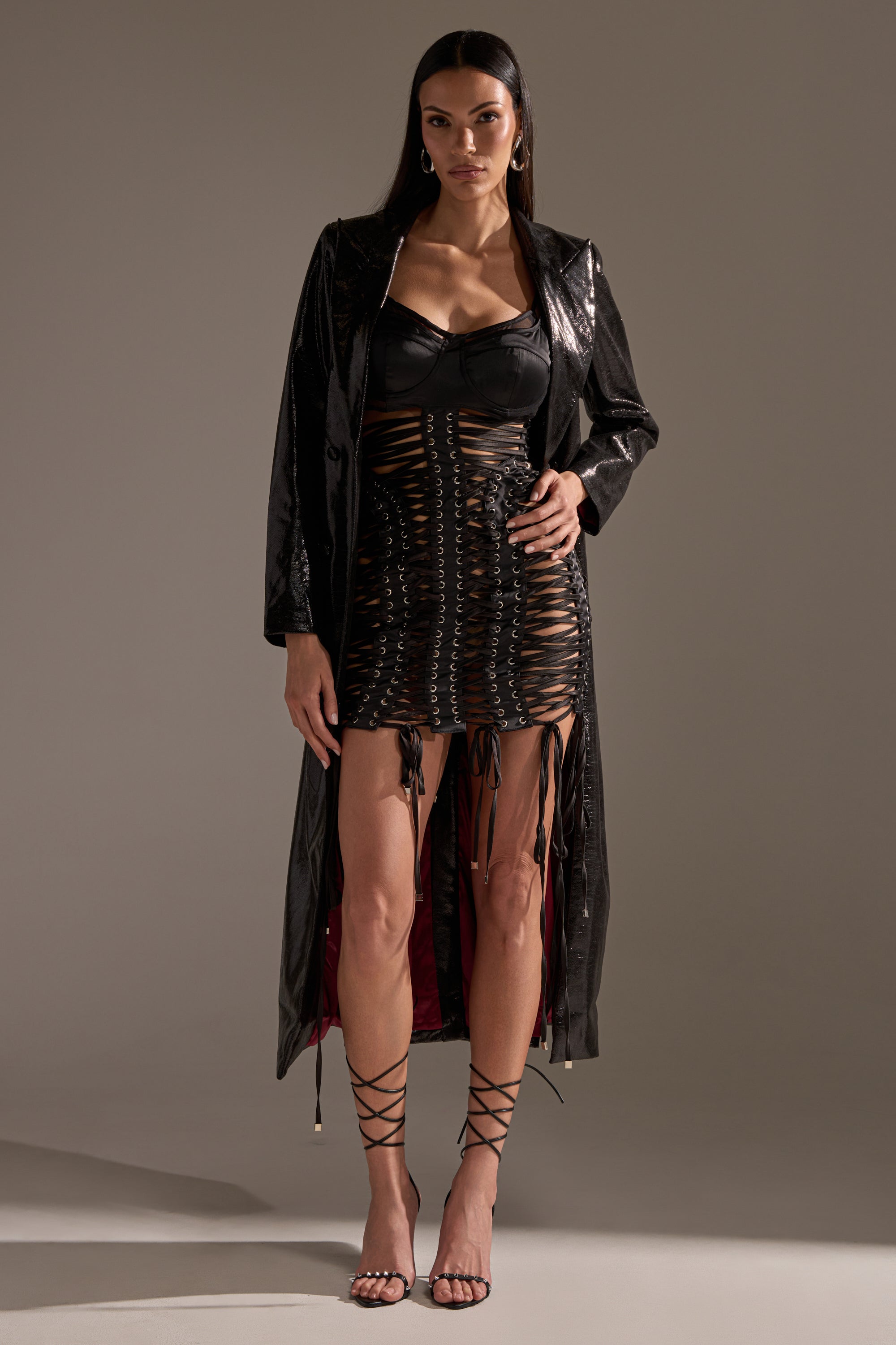 A woman with straight dark hair and a confident look wears the UNBREAKABLE SATIN LACE UP MINI DRESS under a glossy black coat with strappy sandals, standing against a plain background.