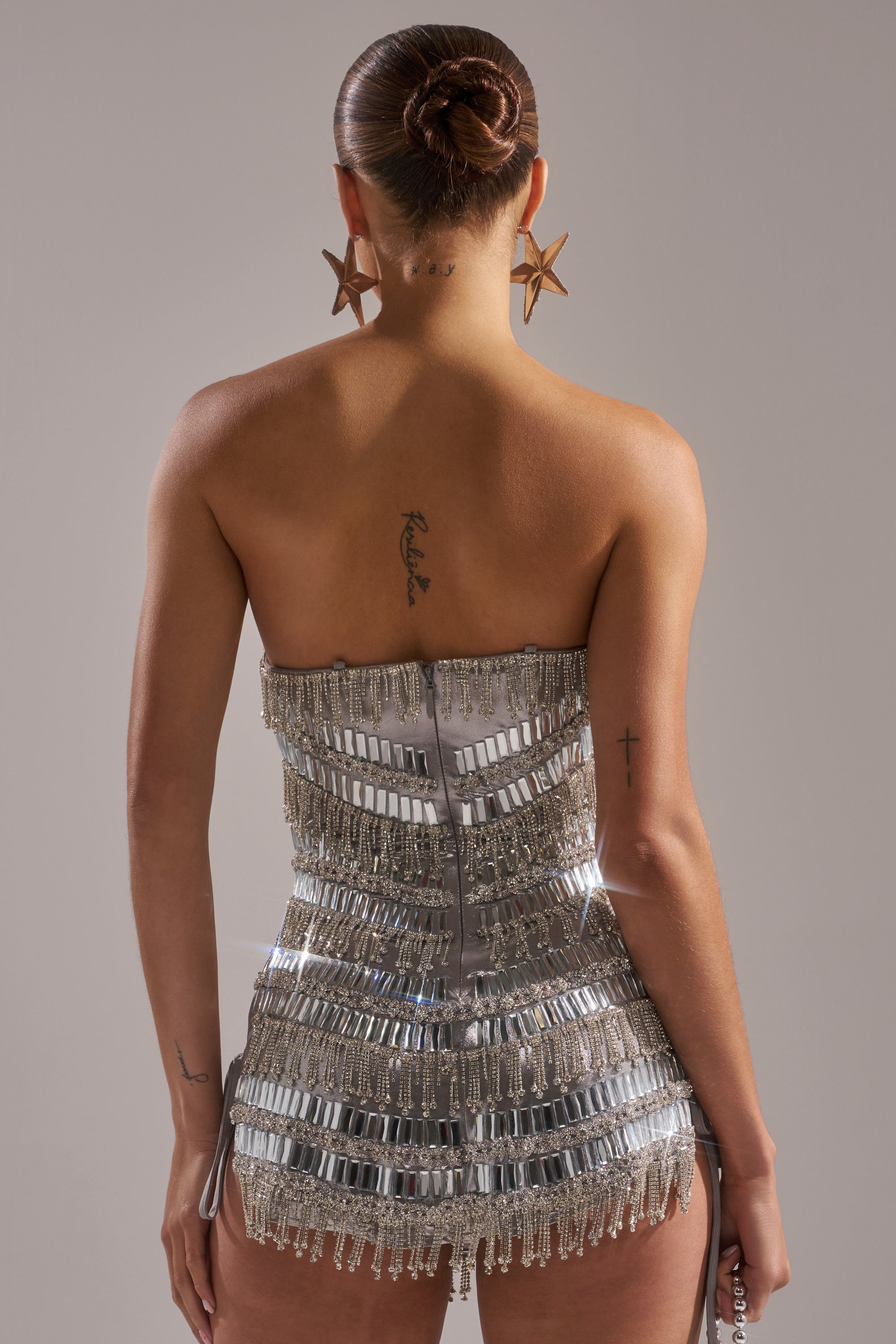 A woman with a sleek bun and large star earrings wears the CHANDELIER RHINESTONE MINI DRESS, a strapless metallic party dress with shimmering fringe, standing with her back to the camera, revealing tattoos on her upper back and arms.