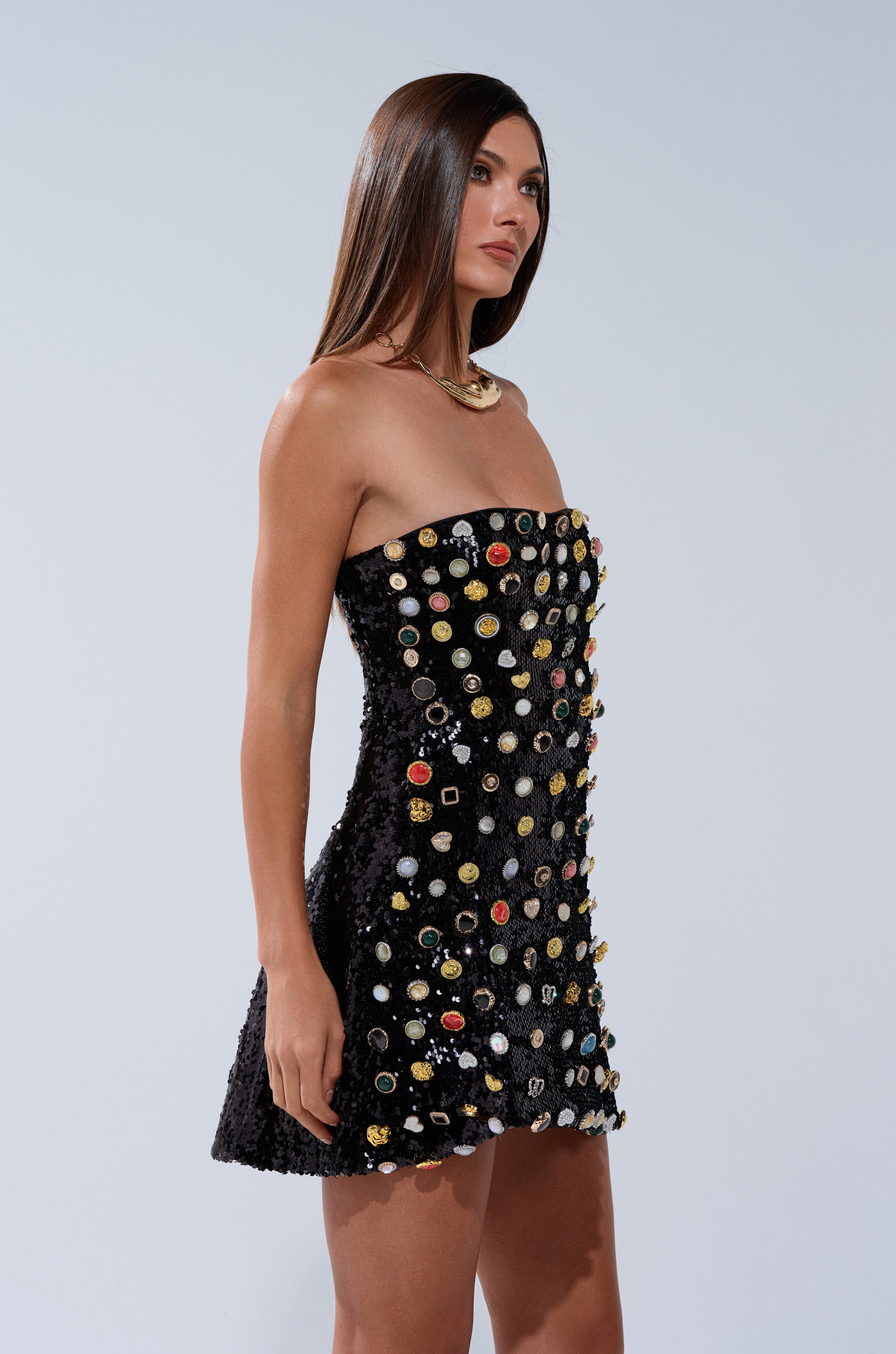 A woman with straight brown hair wears the NEVER NONCHALANT BUTTON MINI DRESS, a strapless, sequin-adorned mini dress, paired with a gold necklace and standing against a plain light background.