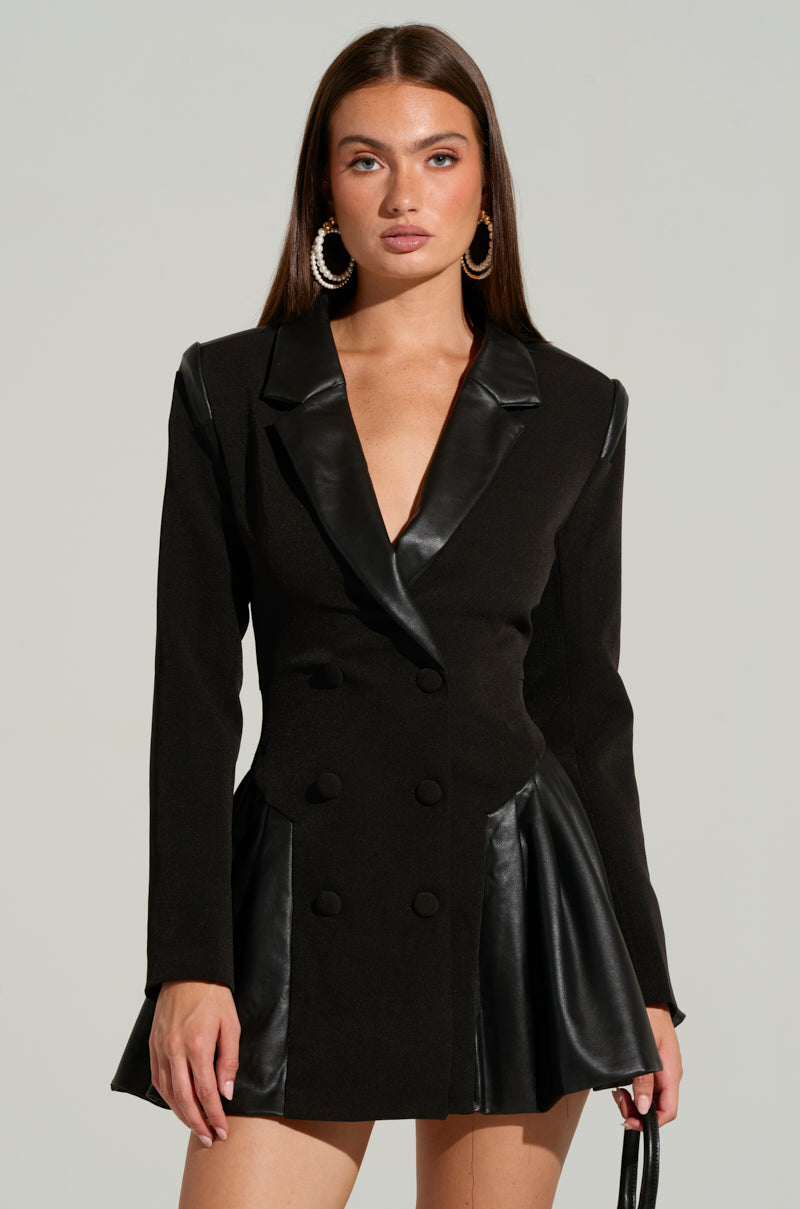 A woman with long, straight hair is wearing the BACK TO BUSINESS FAUX LEATHER DETAIL PLEATED BLAZER DRESS, featuring a black pleated design with double-breasted button front closures and faux leather lapels and skirt details. She has large hoop earrings and is standing against a neutral background, holding a small black handbag by her side.