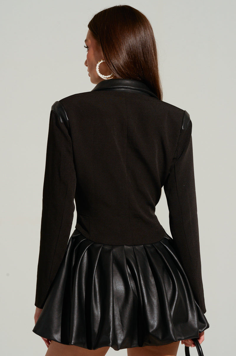 A woman is shown from the back wearing the "BACK TO BUSINESS FAUX LEATHER DETAIL PLEATED BLAZER DRESS." Her straight long hair falls down her back, and she wears large hoop earrings. The background is plain and light-colored.