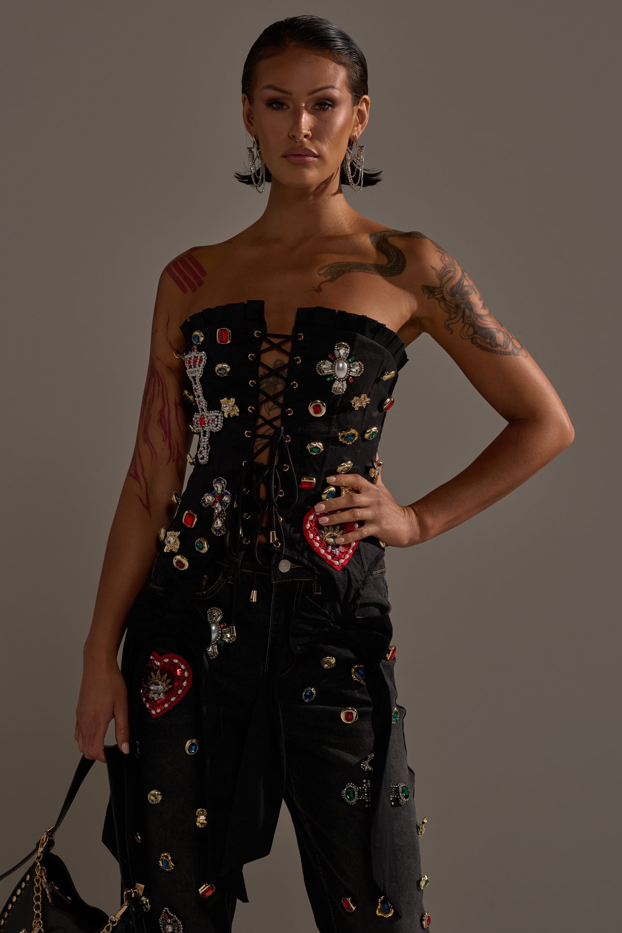 A woman with slicked-back hair and tattoos poses confidently in the MAEVE EMBELLISHED CORSET and jeweled pants, holding a black bag against a neutral background for an avant-garde fashion look. 0