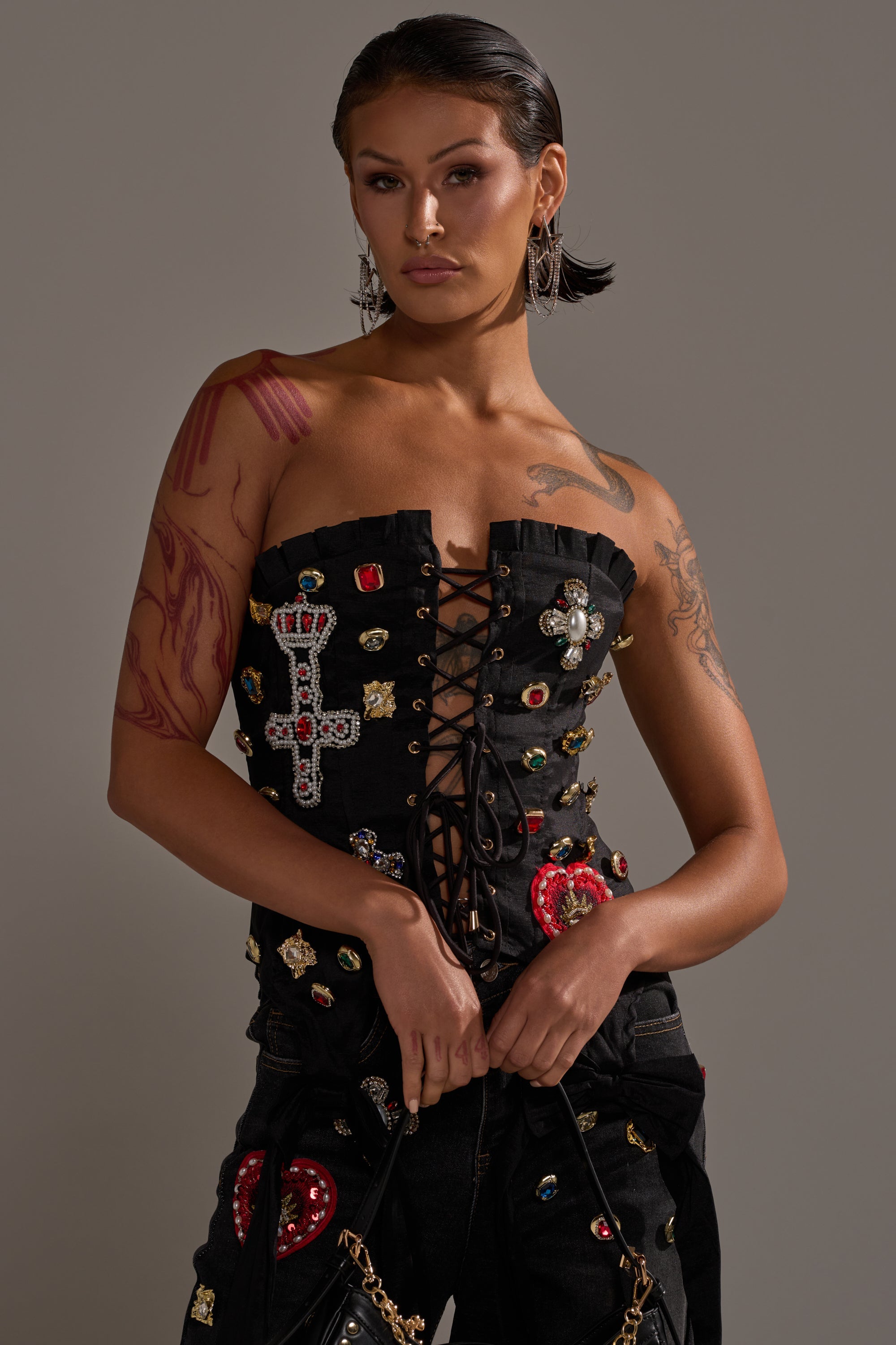 A person with dark hair and tattoos models the MAEVE EMBELLISHED CORSET, featuring colorful gems and a jeweled cross, styled with matching black pants while posing confidently against a neutral background. 1
