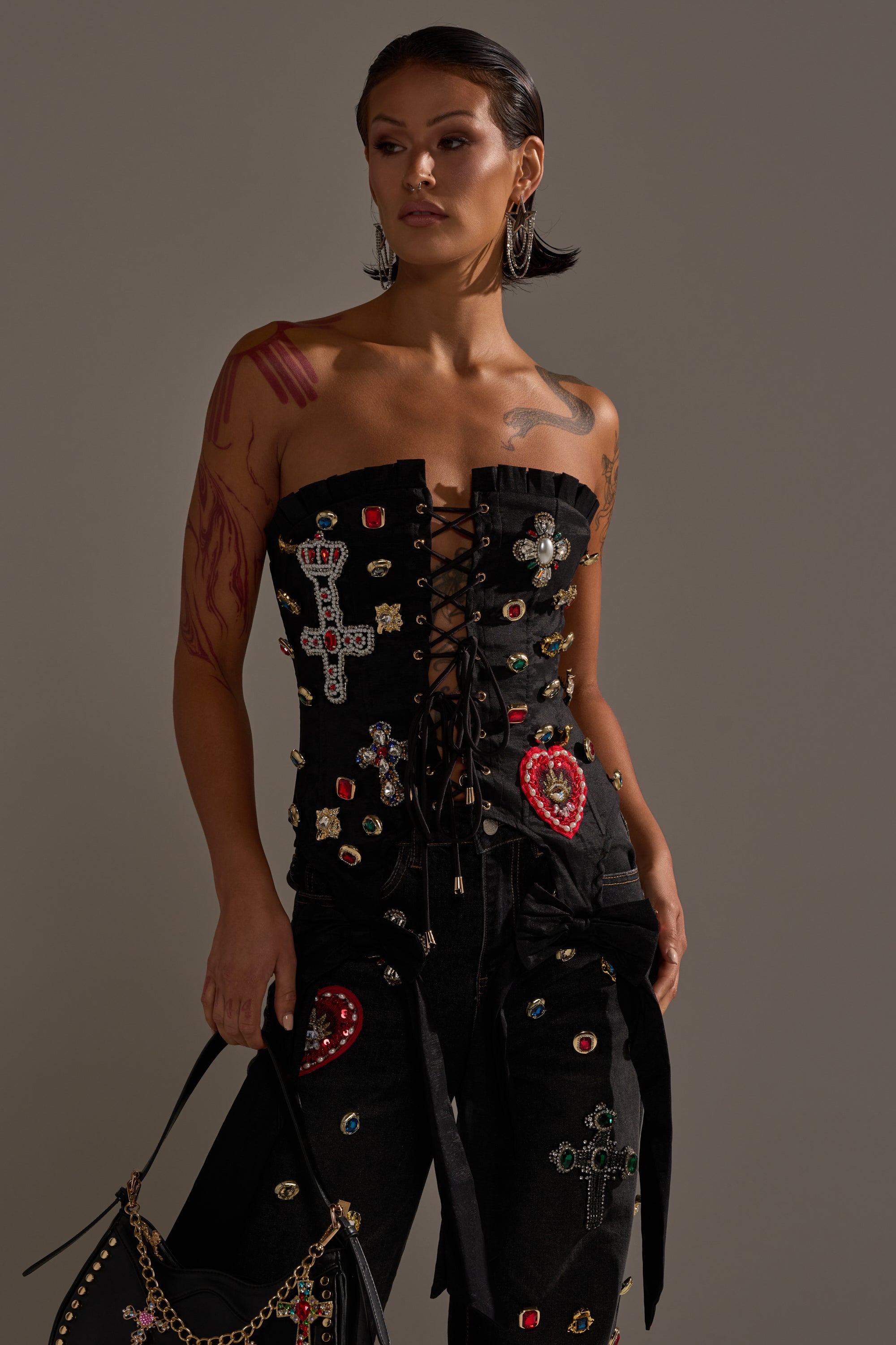 A person with short dark hair wears the MAEVE EMBELLISHED CORSET and pants adorned with jeweled cross and heart details, hoop earrings, and carries a black handbag with a gold chain. 2
