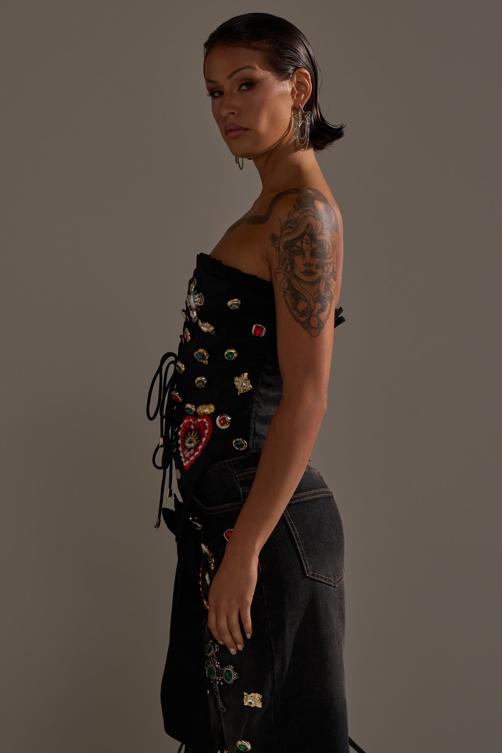 A woman with short dark hair and an arm tattoo wears the MAEVE EMBELLISHED CORSET, a black strapless jeweled corset with a matching skirt, standing sideways against a plain background and looking at the camera. 3