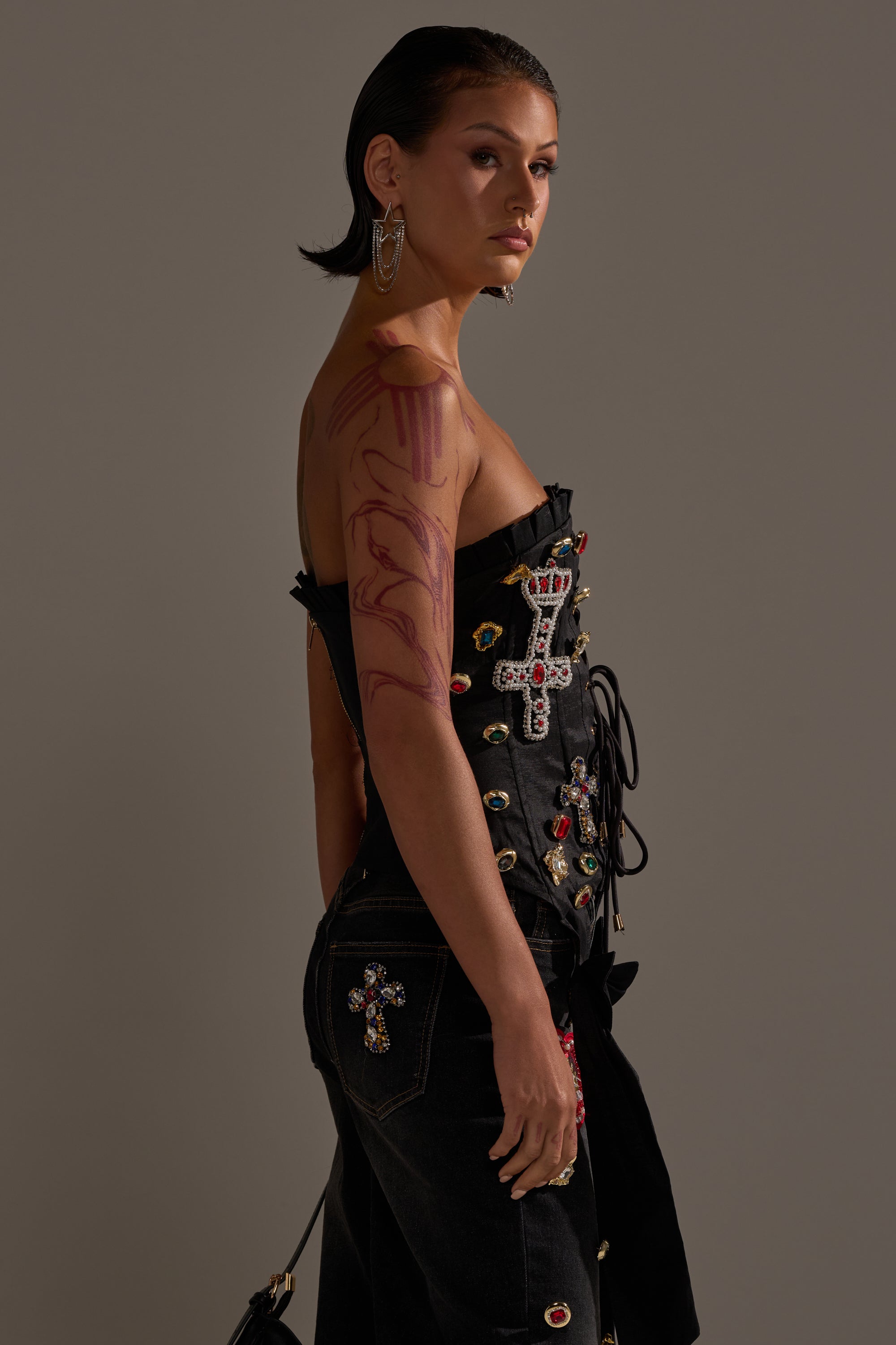 Someone with slicked-back hair wears the MAEVE EMBELLISHED CORSET and matching jewel-adorned jeans, featuring colorful crosses, bold earrings, and visible red body art while posing against a neutral background. 4