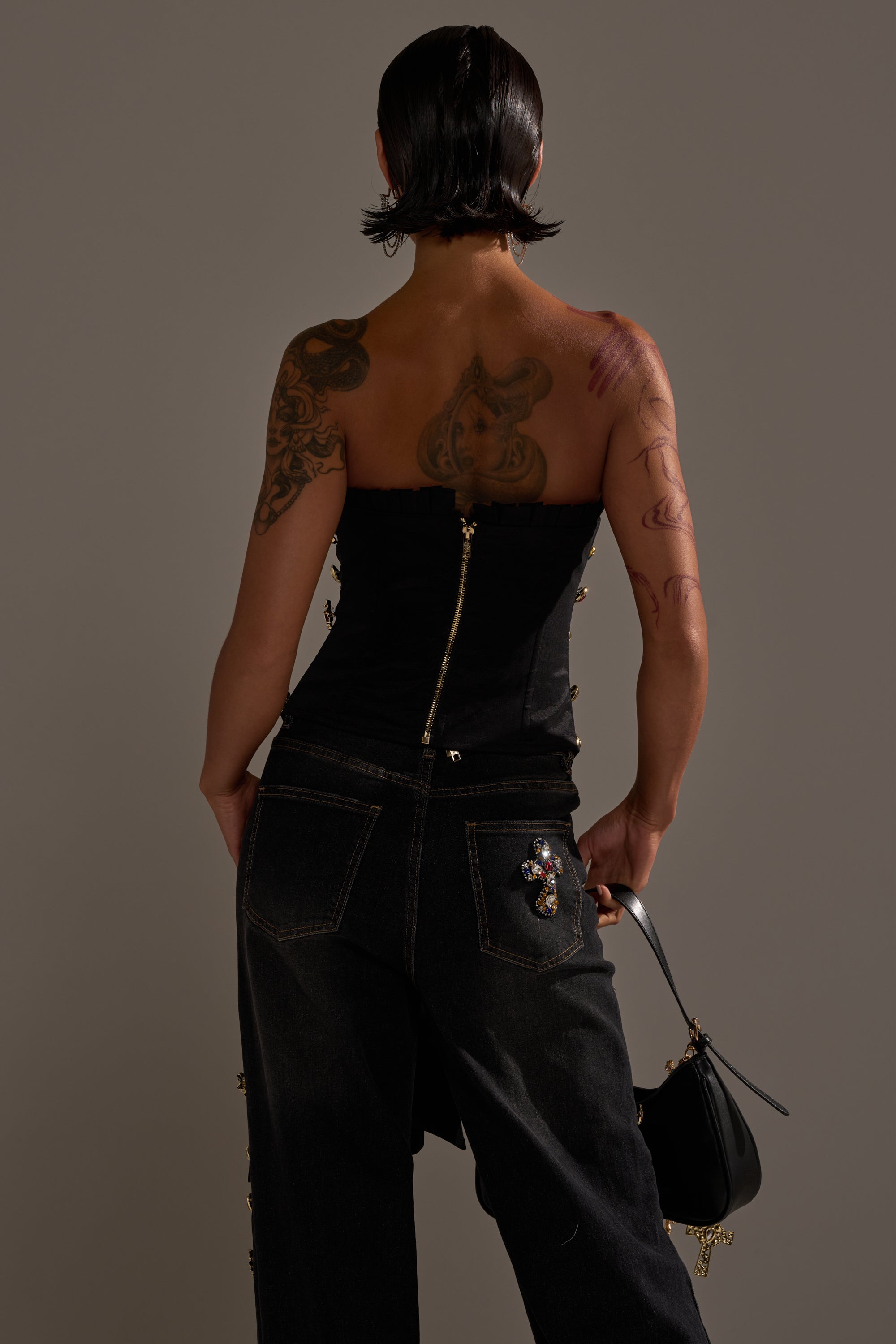 A person with short dark hair and tattoos wears the MAEVE EMBELLISHED CORSET, dark pants with a jeweled patch, and holds a small black handbag while facing away against a plain gray background. 5