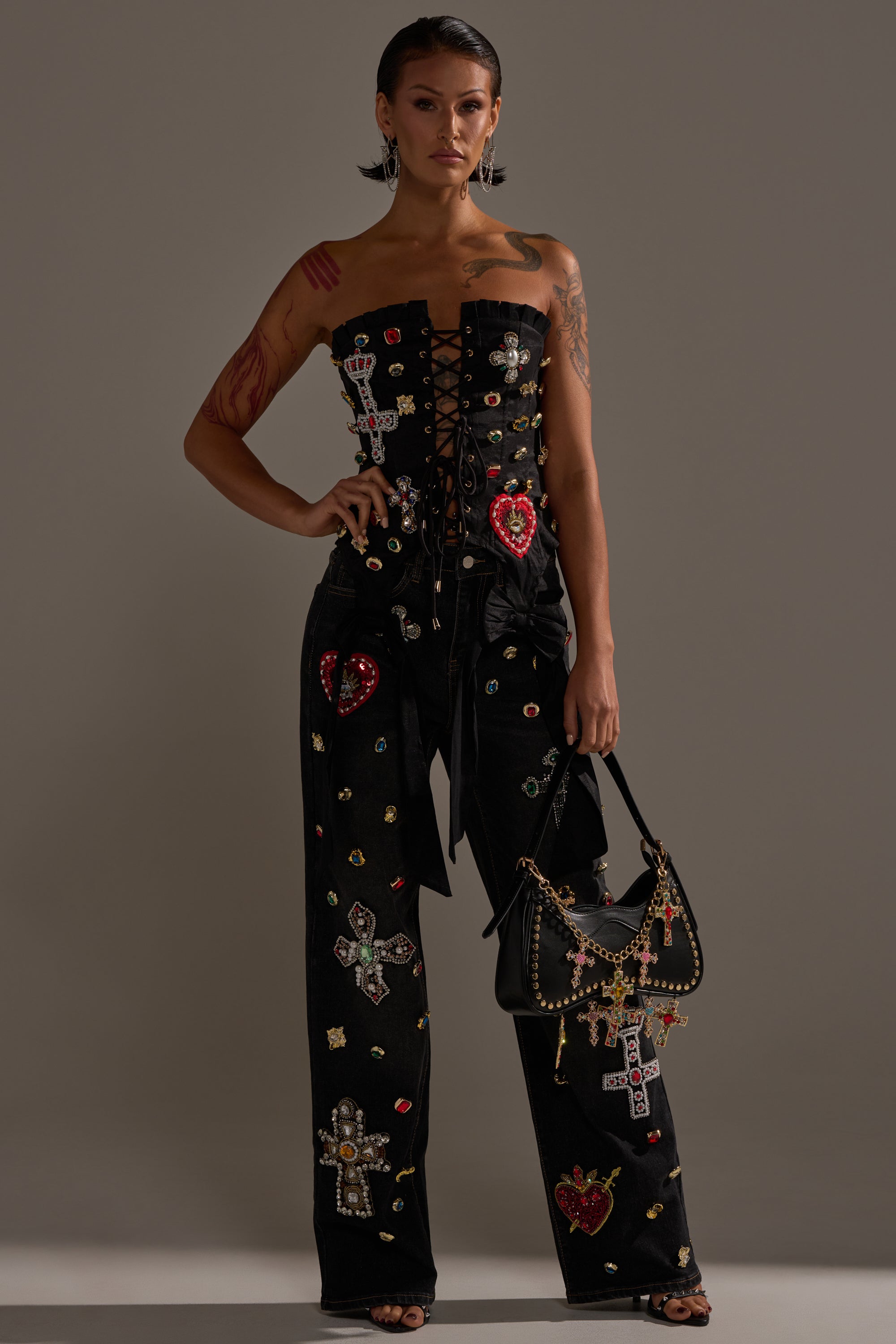 A person stands wearing the MAEVE EMBELLISHED CORSET—strapless and decorated with colorful jeweled patches and cross motifs—paired with matching pants, and holds a black handbag featuring gold chain accents. 6