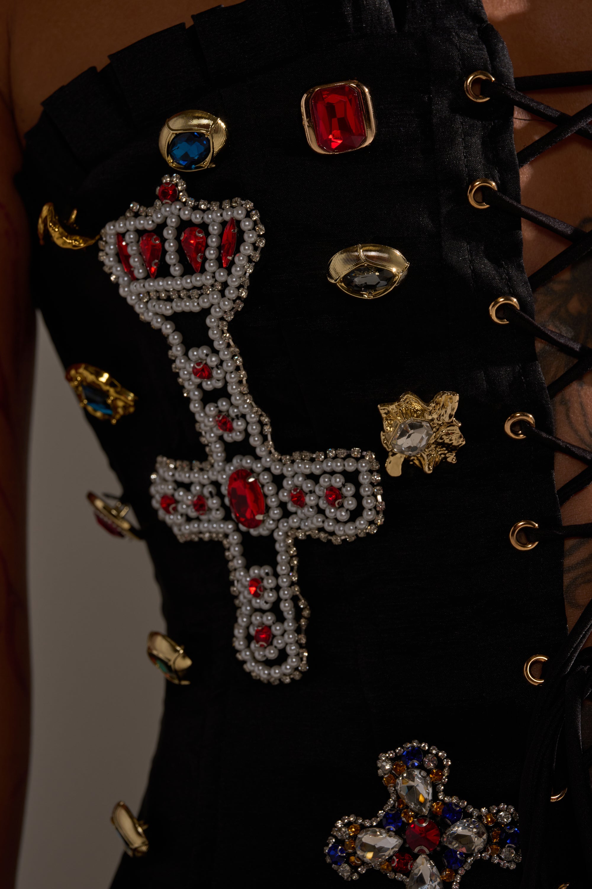 Close-up of the MAEVE EMBELLISHED CORSET, adorned with black laces, jeweled and beaded details, a bold beaded cross with red and white gems, and gold accents—a striking choice for avant-garde fashion fans. 8