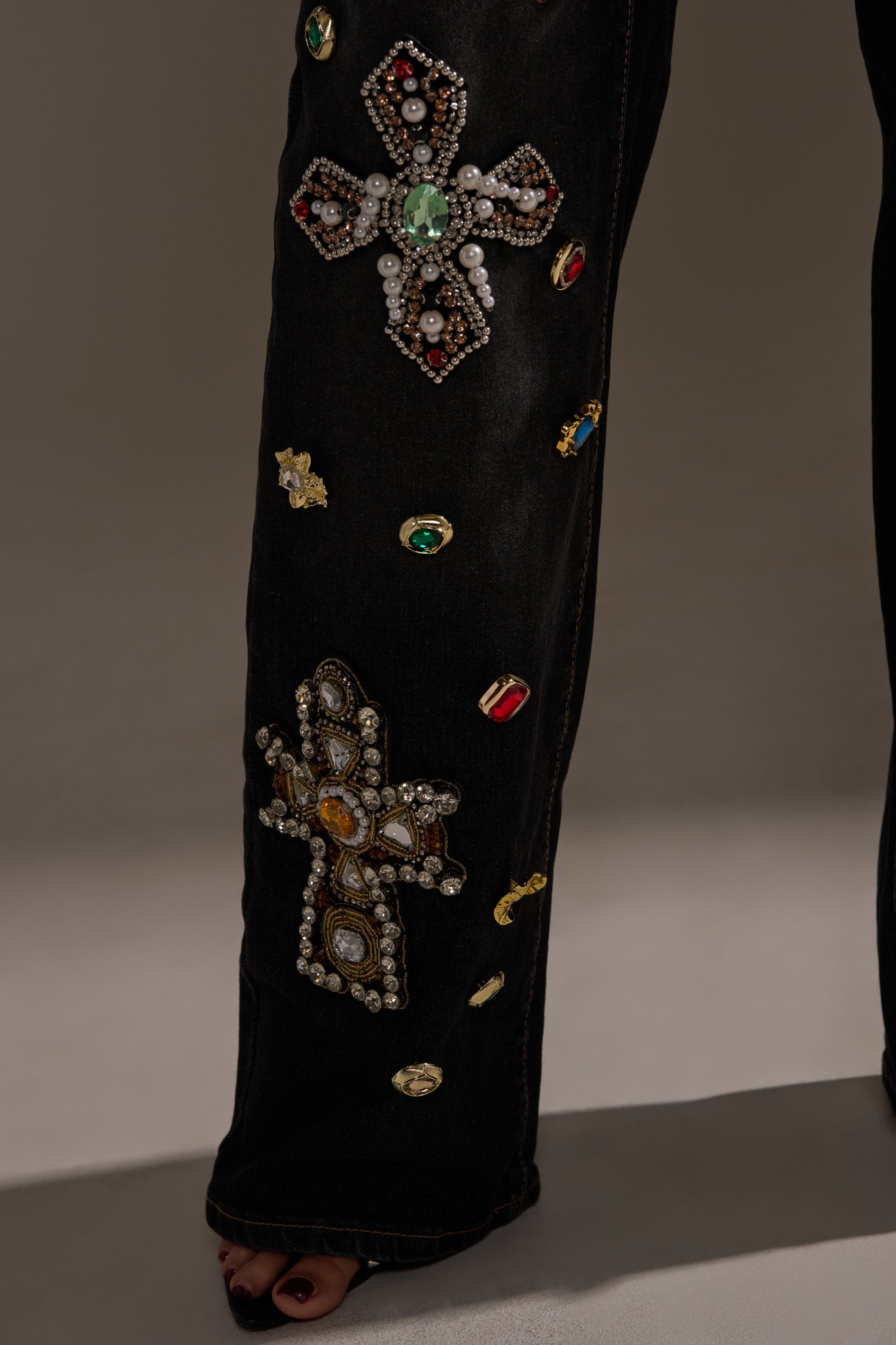 A close-up of the FEELING EXTRA BLESSED EMBELLISHED DENIM shows a high-waisted black jean leg adorned with large jeweled crosses and ovals. The wearer stands barefoot against a neutral background.