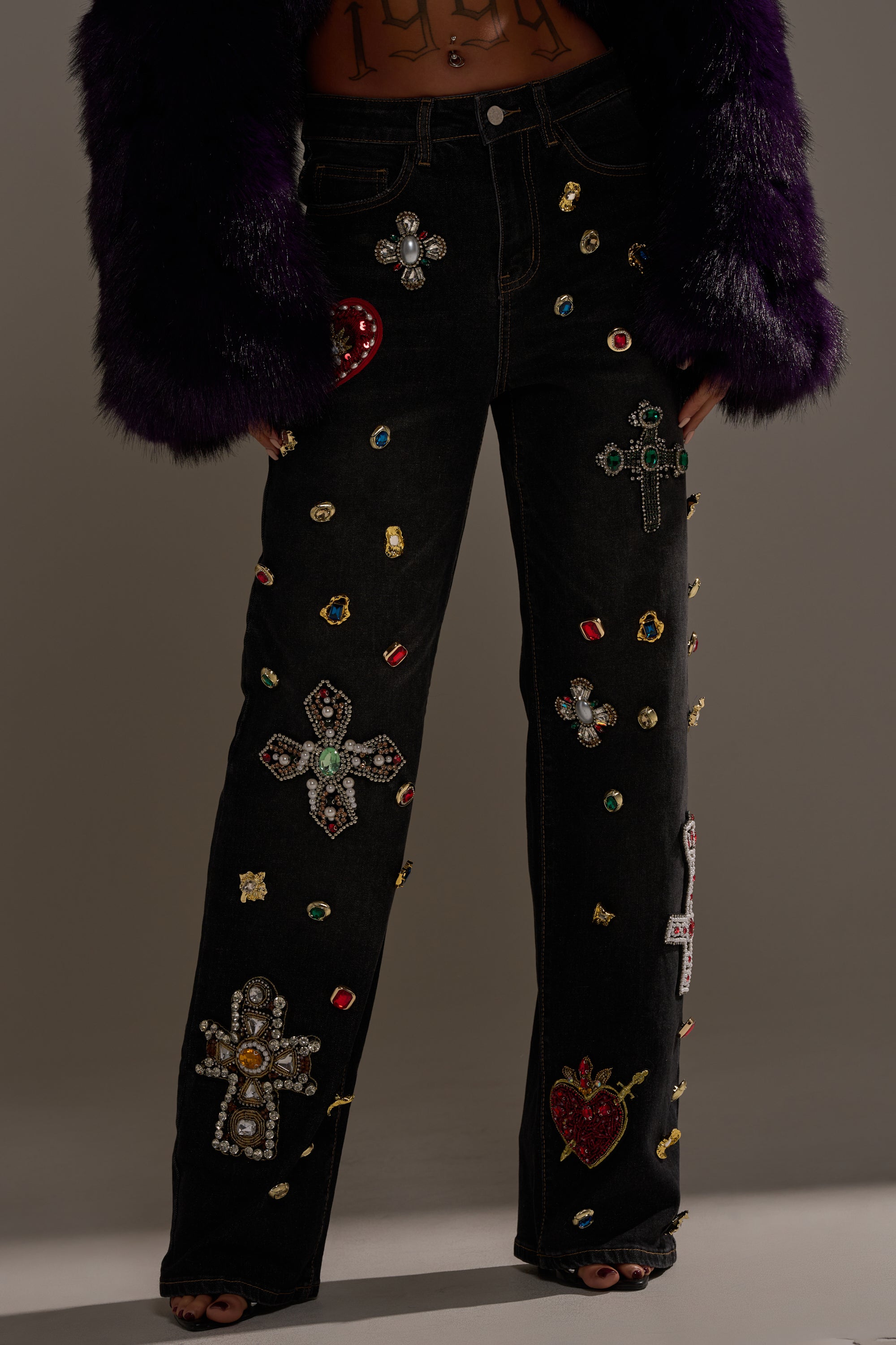 Wearing the FEELING EXTRA BLESSED EMBELLISHED DENIM—high-waisted black jeans with jeweled crosses and heart patches—paired with a dark purple faux fur jacket. Only the lower torso and legs are visible.