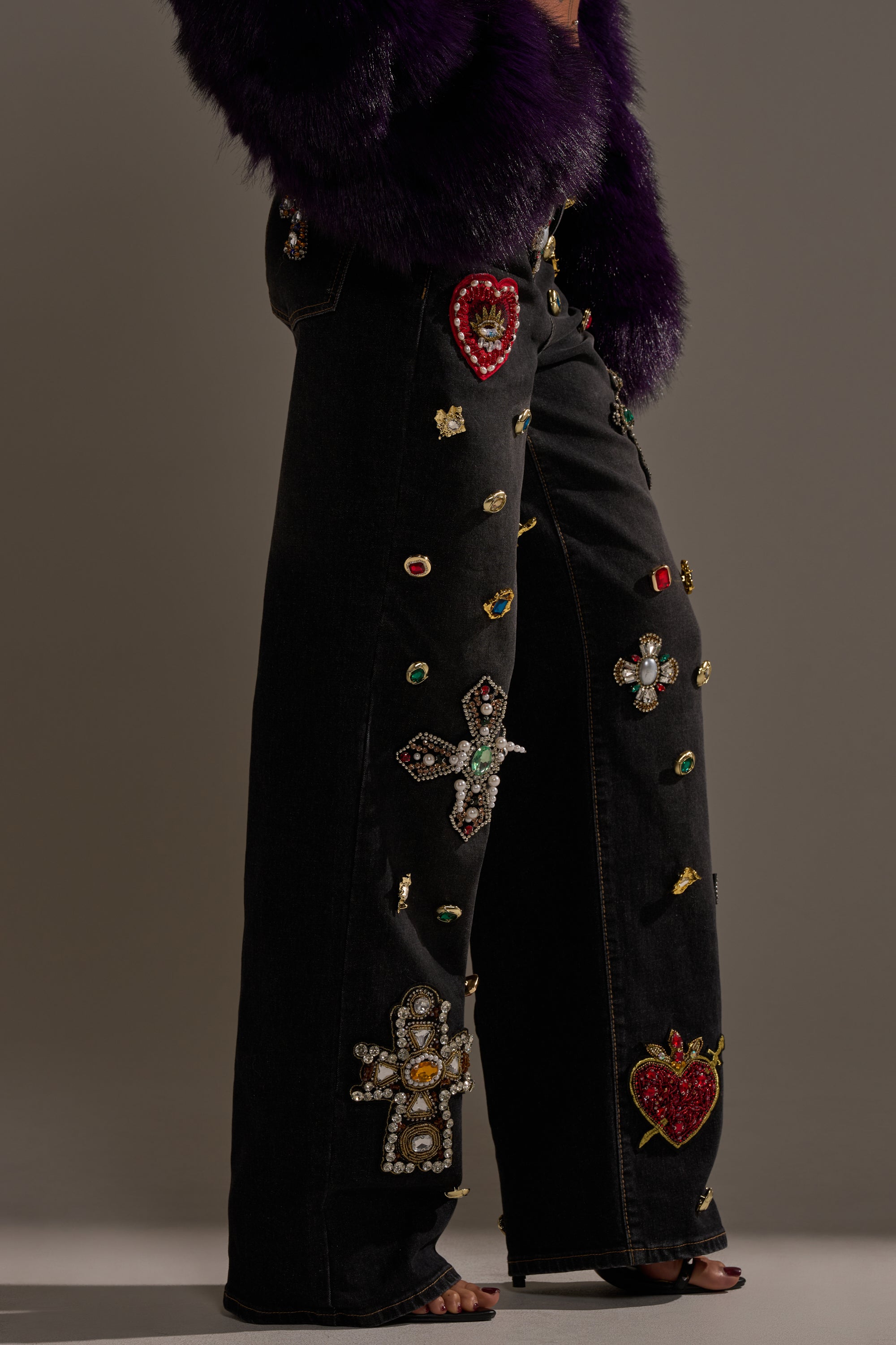 Wearing the FEELING EXTRA BLESSED EMBELLISHED DENIM—high-waisted black jeans with jeweled hearts, crosses, and flowers—styled with a deep purple faux fur coat and open-toe heels for a bold, statement look.