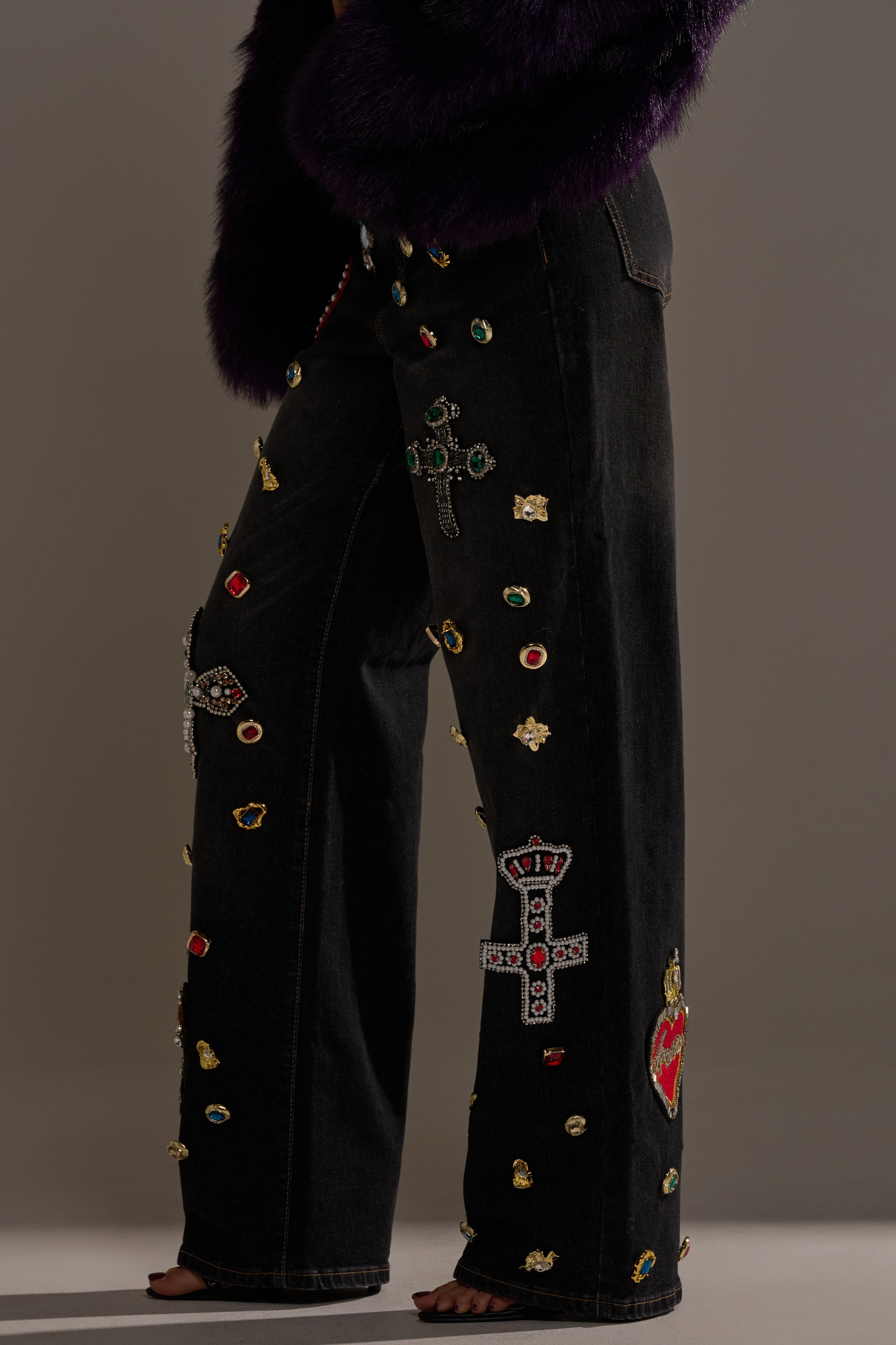 Wearing the FEELING EXTRA BLESSED EMBELLISHED DENIM jeweled jeans with colorful gem and cross-shaped accents and a dark purple textured jacket, a barefoot individual stands against a neutral background.