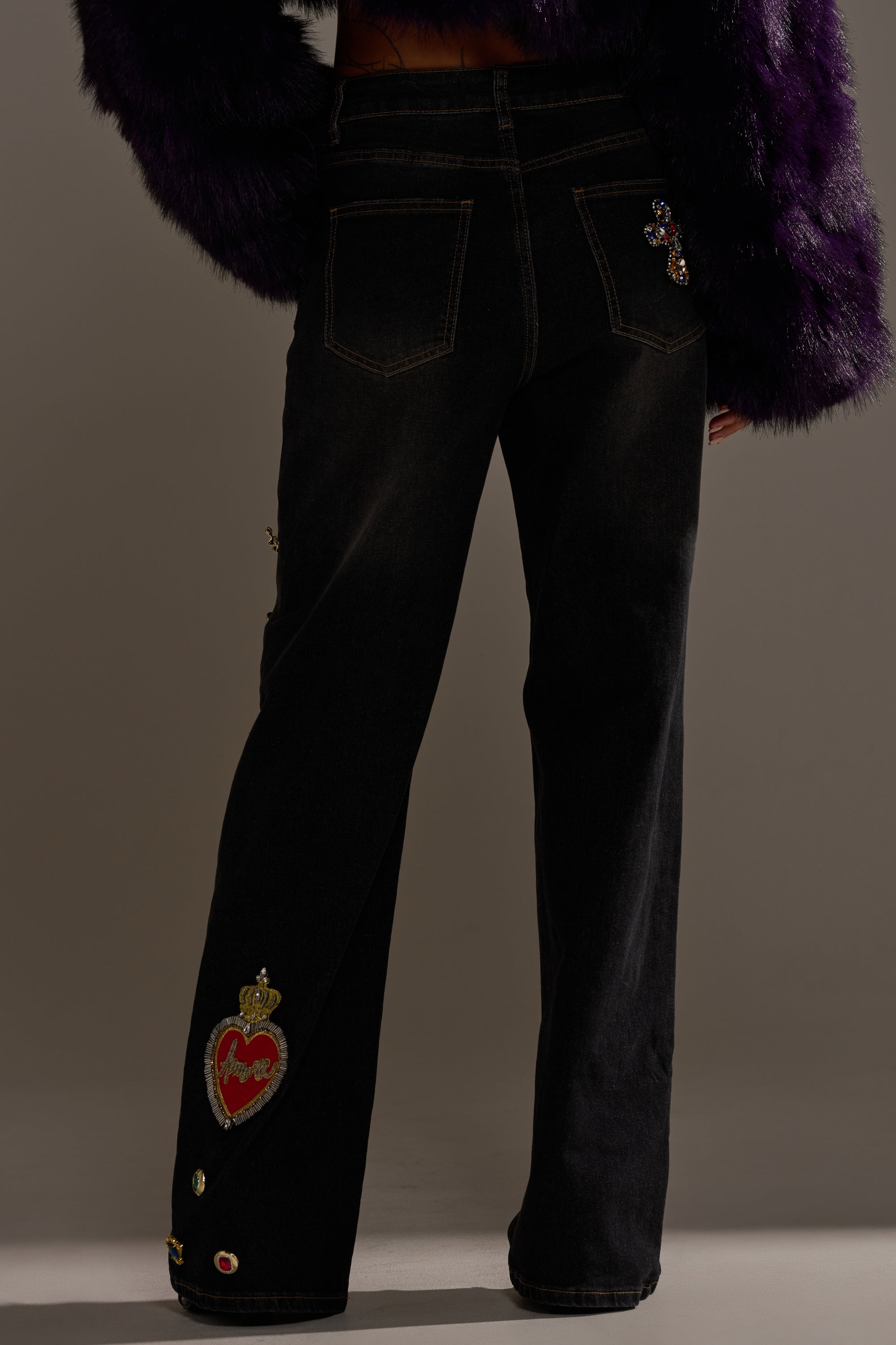 A person wears FEELING EXTRA BLESSED EMBELLISHED DENIM—high-waisted black jeans with decorative patches, like a heart near the left ankle—paired with a dark purple fuzzy jacket. Photo is from behind, showcasing the denim’s embellishments.