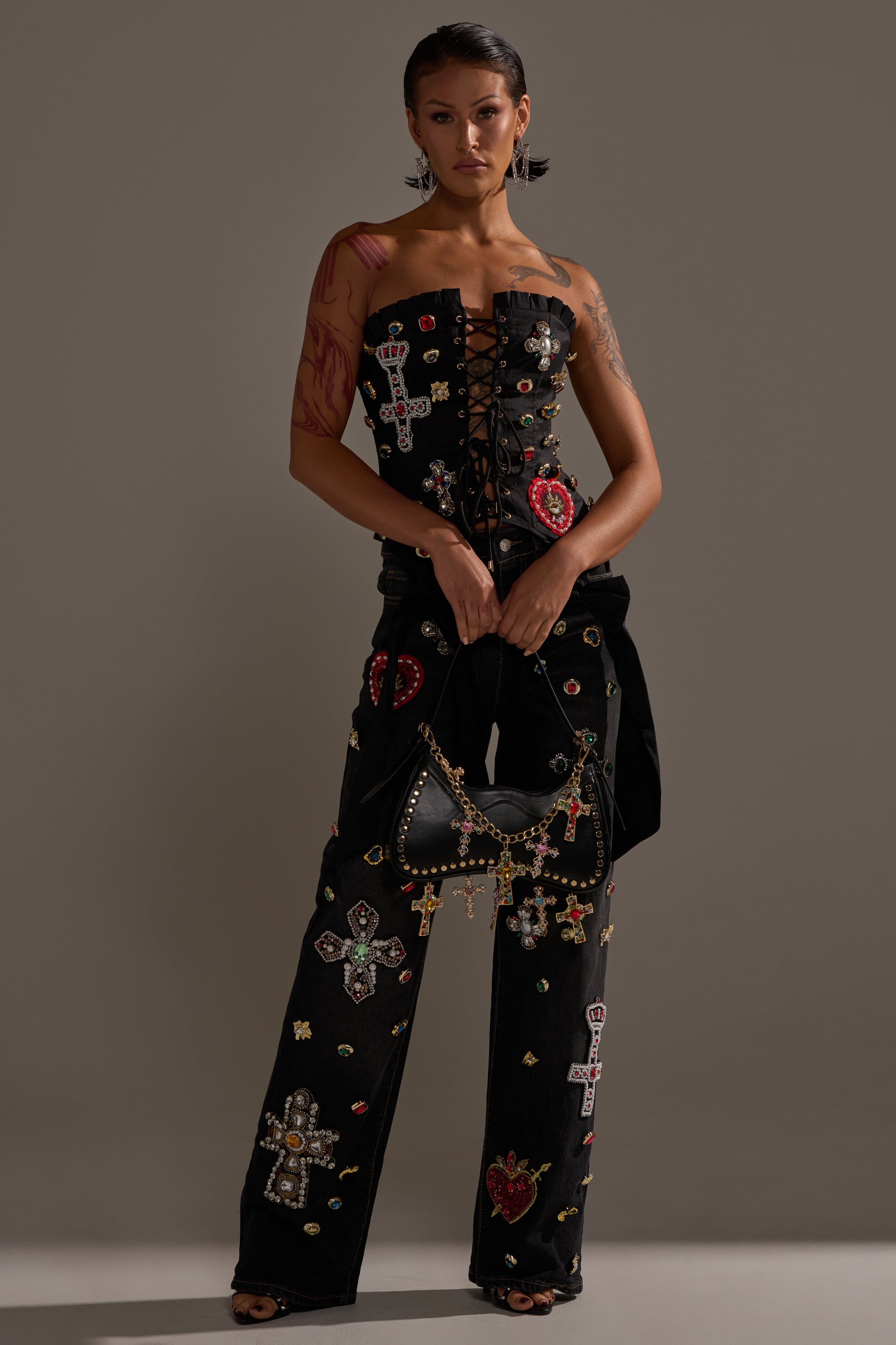 A woman faces forward in FEELING EXTRA BLESSED EMBELLISHED DENIM—high-waisted black jeans with jeweled cross and heart details—paired with a black strapless lace-up top, holding a black bag against a neutral gray background.