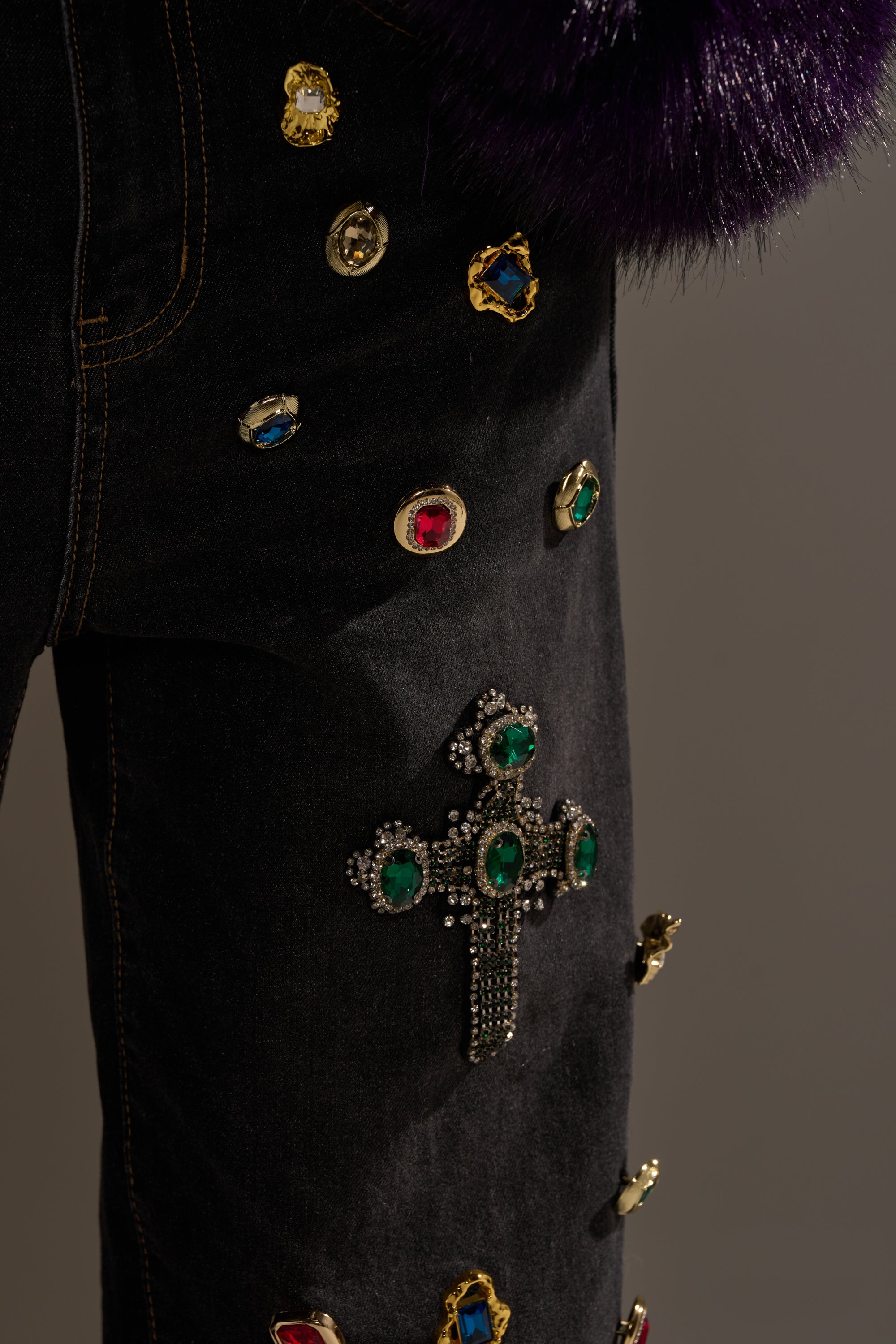 Close-up of FEELING EXTRA BLESSED EMBELLISHED DENIM, high-waisted black jeans with vibrant gemstone accents and a jeweled cross, styled with a dark purple faux fur piece for a bold statement look.