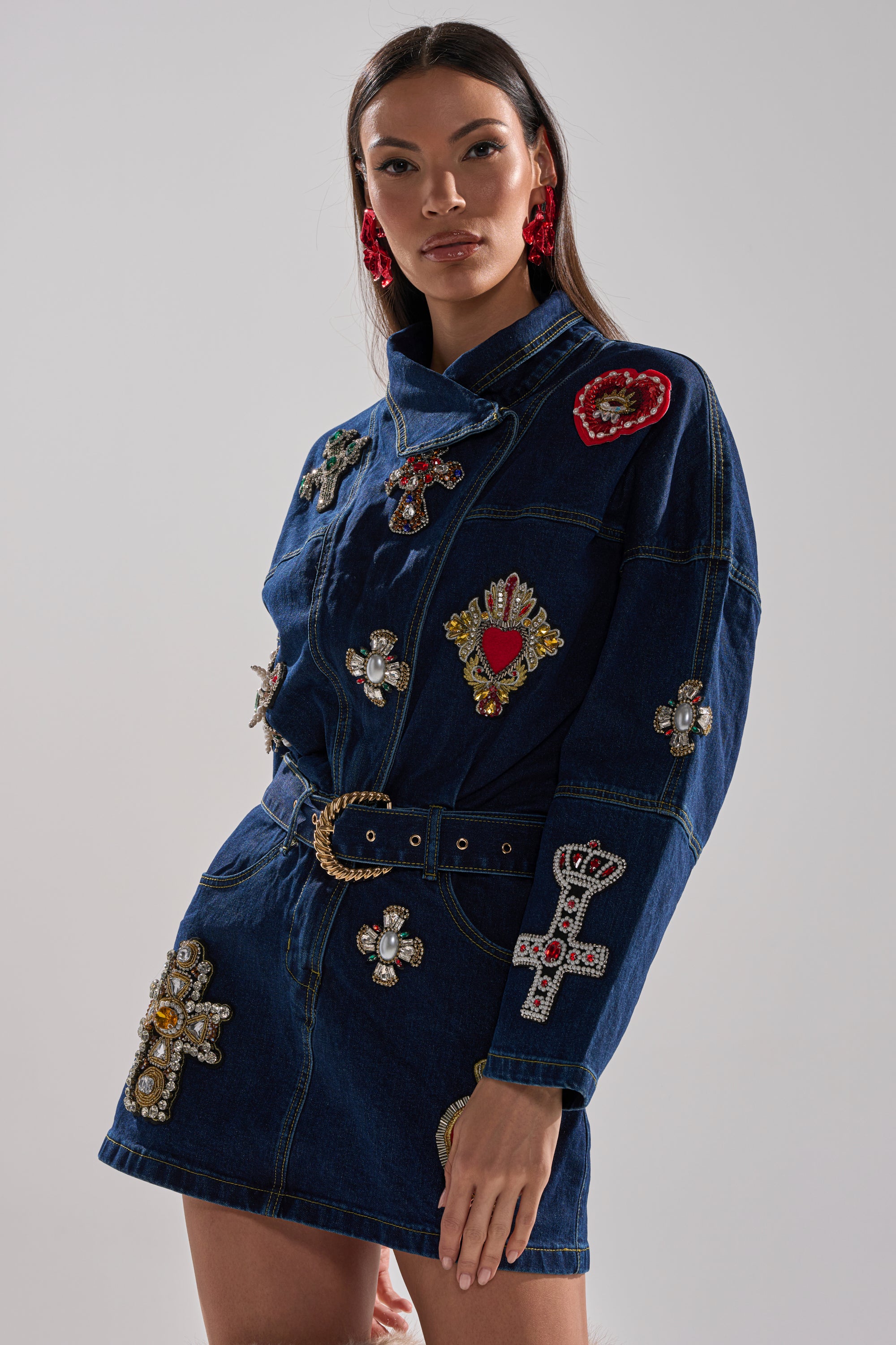 Wearing the PRETTIEST PROBLEM DENIM PATCHWORK MINI DRESS, featuring a belted waist, bejeweled cross and heart patches, and red statement earrings, a woman stands against a plain light gray background.