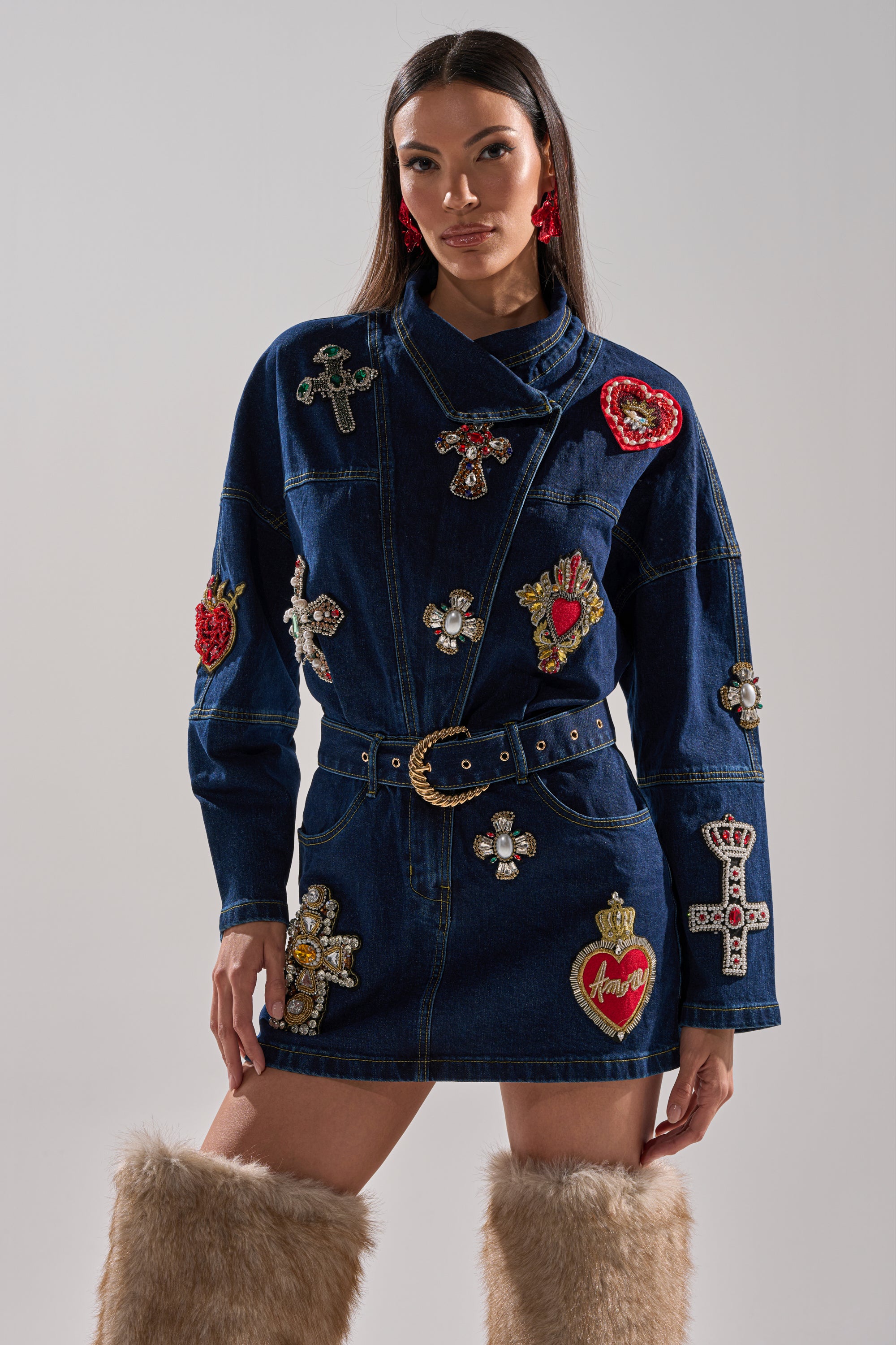 A woman wears the PRETTIEST PROBLEM DENIM PATCHWORK MINI DRESS with bejeweled crosses and hearts, a belted waist with gold buckle, red earrings, and tan fur knee-high boots, standing against a plain background.