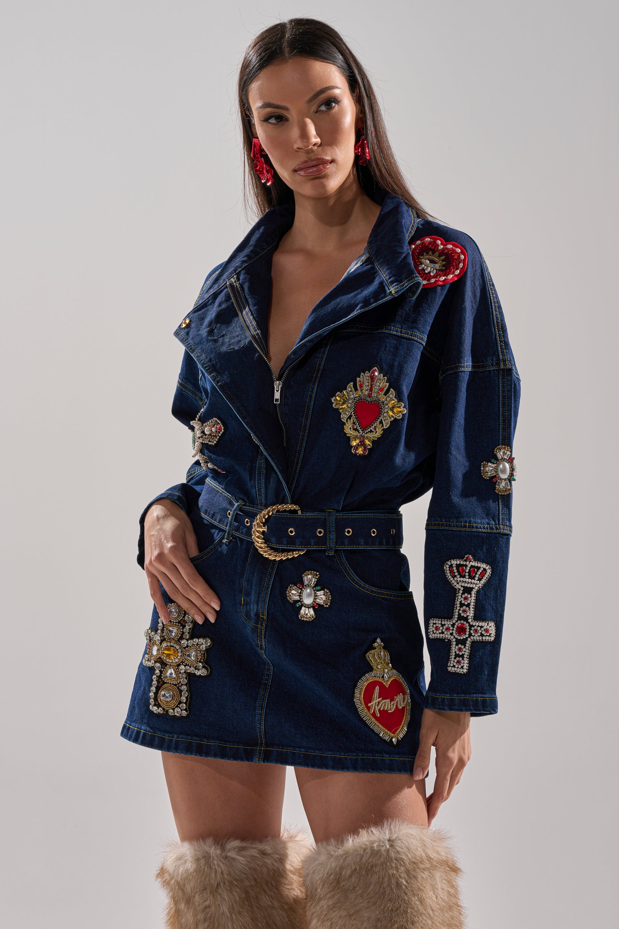Wearing the PRETTIEST PROBLEM DENIM PATCHWORK MINI DRESS with a belted waist, colorful embroidered hearts, and bejeweled crosses, she pairs it with large red earrings and tan furry boots against a plain light background.