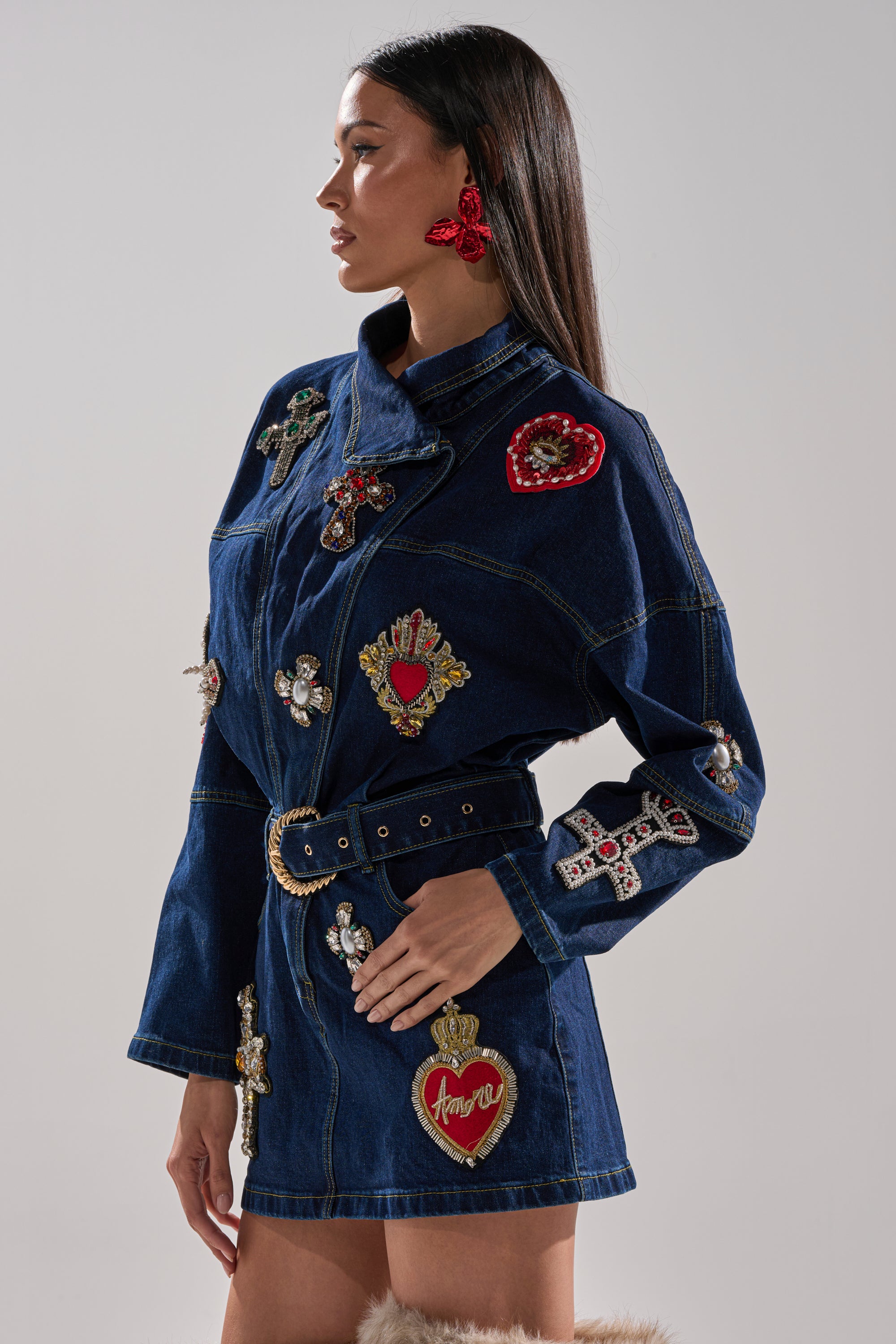 A woman with long dark hair stands in profile against a neutral background, wearing the PRETTIEST PROBLEM DENIM PATCHWORK MINI DRESS featuring a belted waist and jeweled heart and cross appliqués.