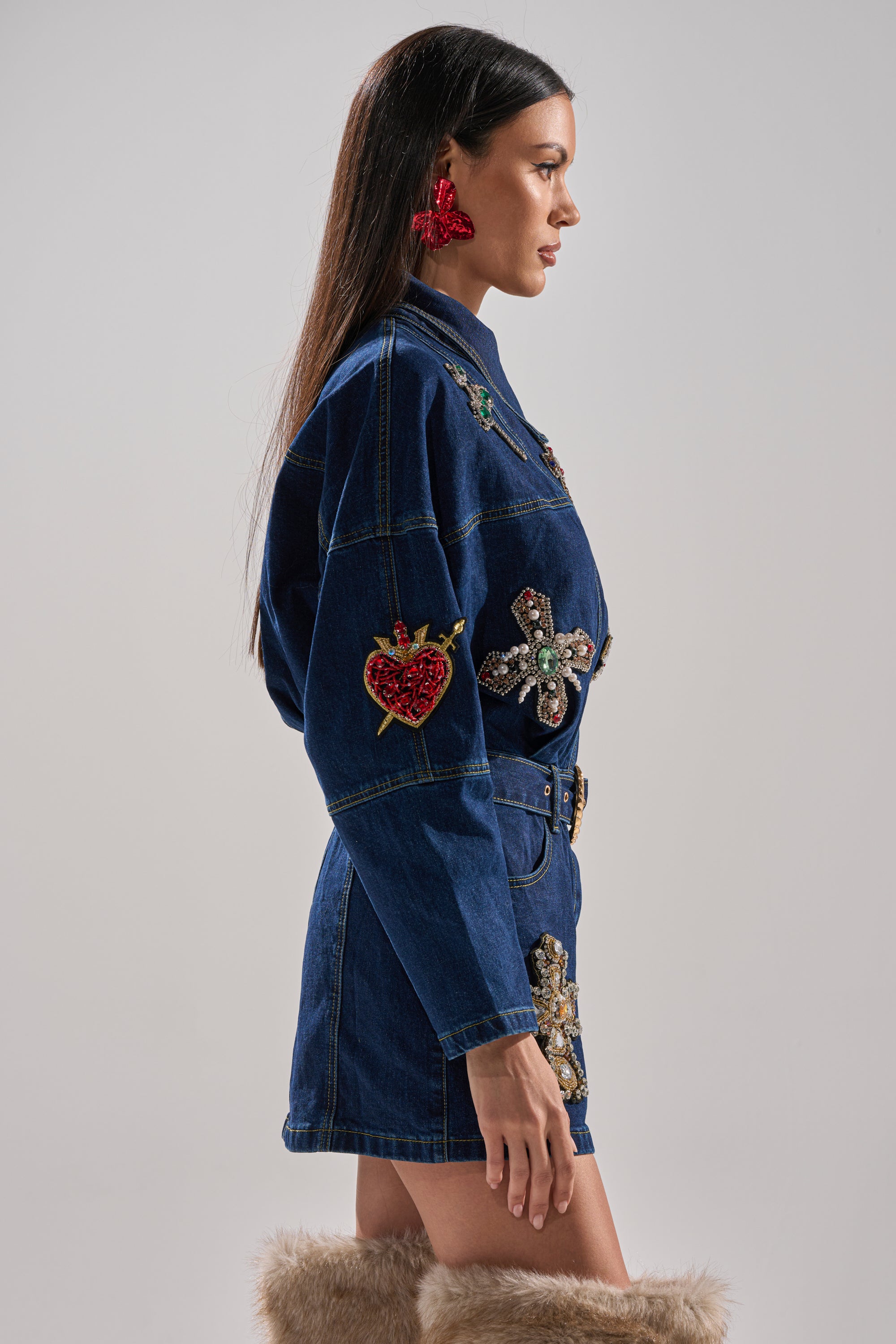 A woman stands in profile against a plain background wearing the PRETTIEST PROBLEM DENIM PATCHWORK MINI DRESS with a belted waist, bejeweled crosses, large red earrings, and tall furry boots.