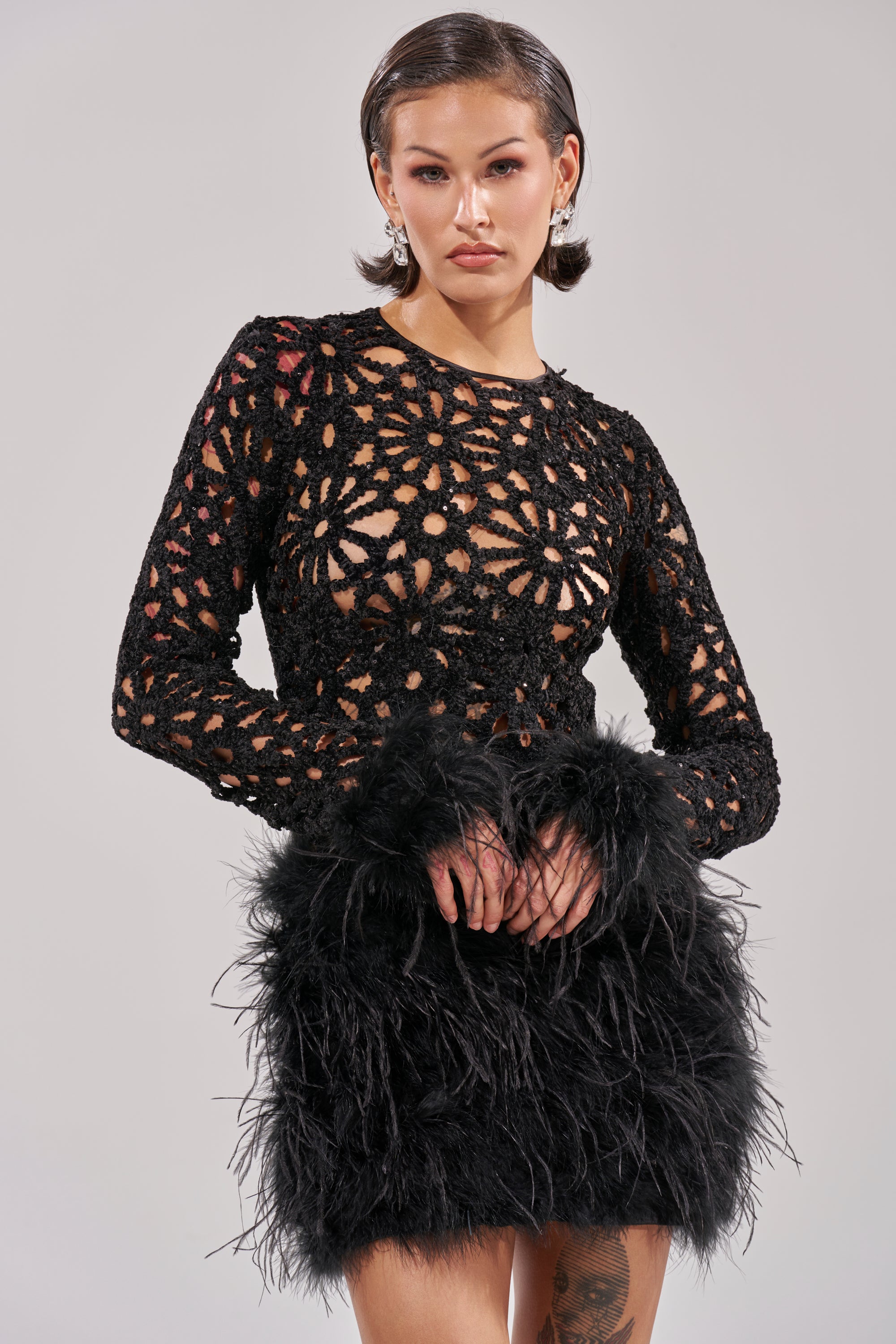 A woman poses in the WHO ME? Knit Crochet Mini Dress with Feathers, featuring a sheer floral lace top, long sleeves, and a textured feathered skirt. Her short, slicked-back hair and neutral makeup complete the look against a plain background.