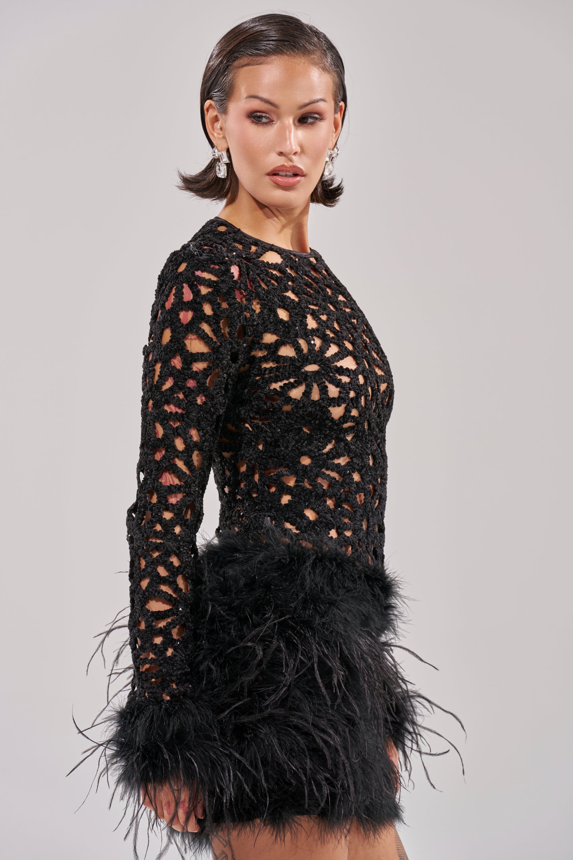 A woman with slicked-back short hair models the WHO ME? Knit Crochet Mini Dress With Feathers—featuring long sleeves, semi-sheer fabric, floral cutouts, and feather trim at the cuffs and hem—posing confidently against a plain light background.