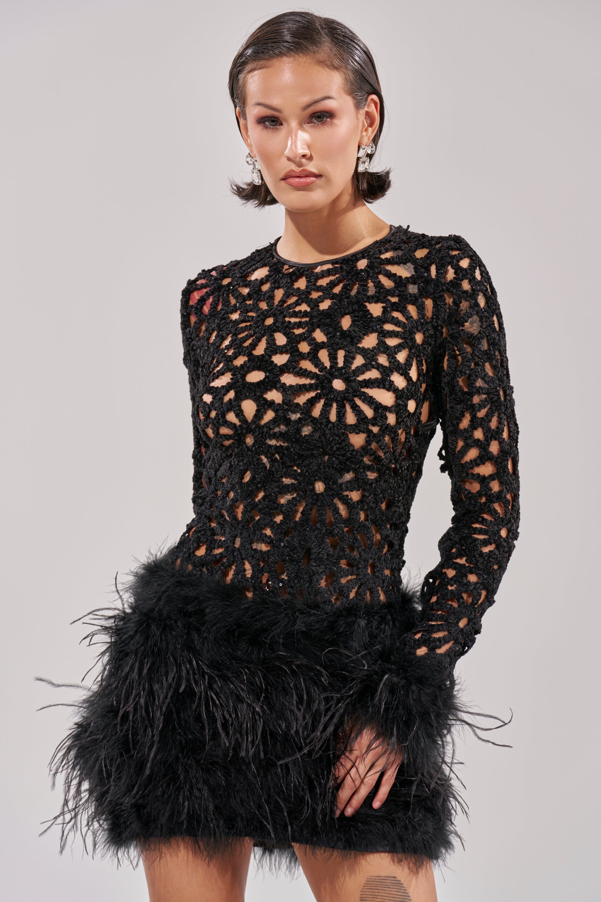 A person wears the WHO ME? KNIT CROCHET MINI DRESS WITH FEATHERS, a long-sleeved black cocktail dress with geometric crochet cutouts on top and a feather-trimmed skirt, standing against a plain light background.