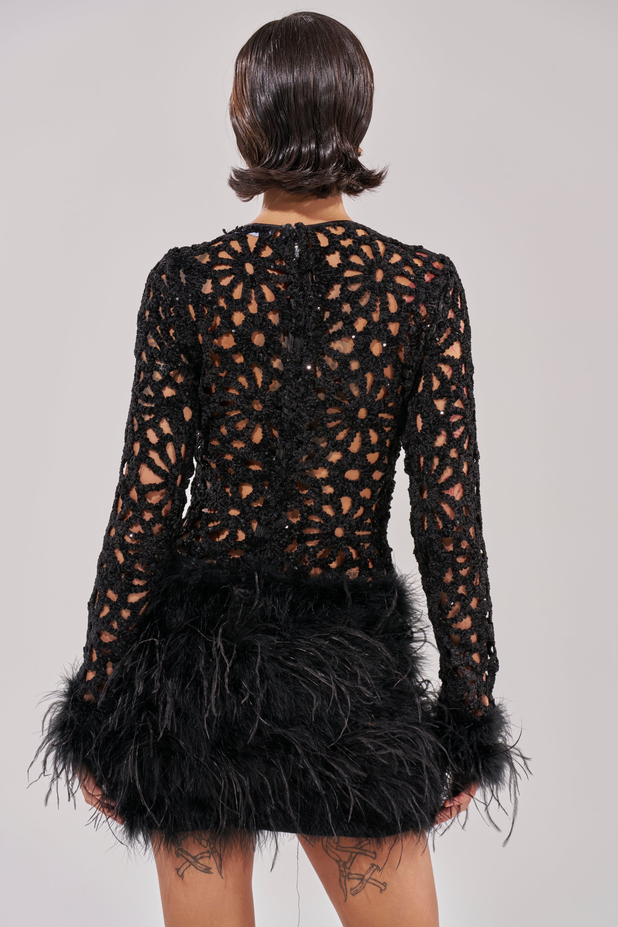 A person with short, dark, wavy hair is shown from behind wearing the WHO ME? KNIT CROCHET MINI DRESS WITH FEATHERS. Their tattooed legs stand out against a plain light gray background.