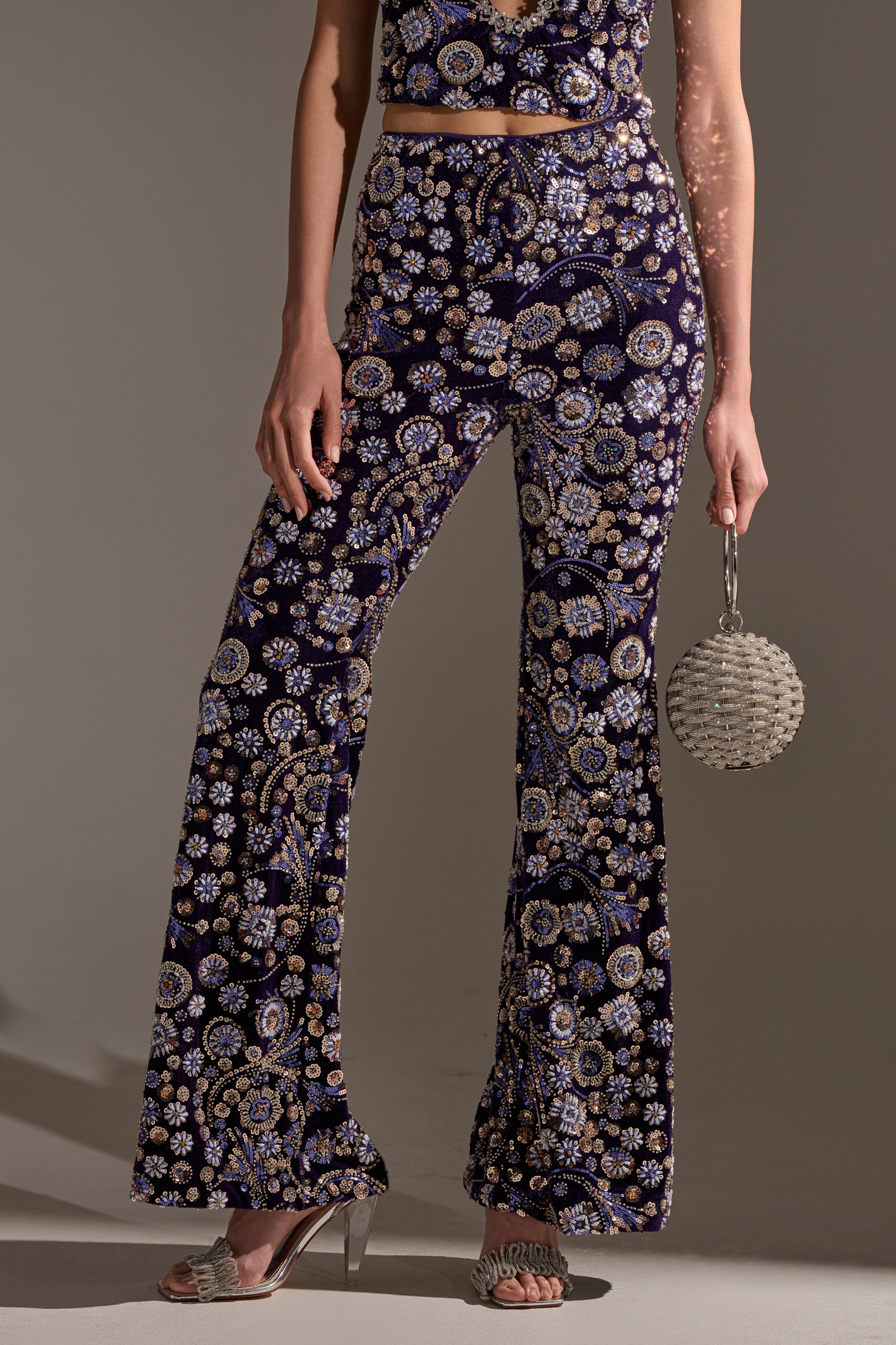 Wearing the QUEEN OF EVERYTHING RHINESTONE TROUSER, a high-rise flared pant in dark purple with floral rhinestone detailing, paired with a matching crop top, a round beaded handbag, and embellished open-toe heels.