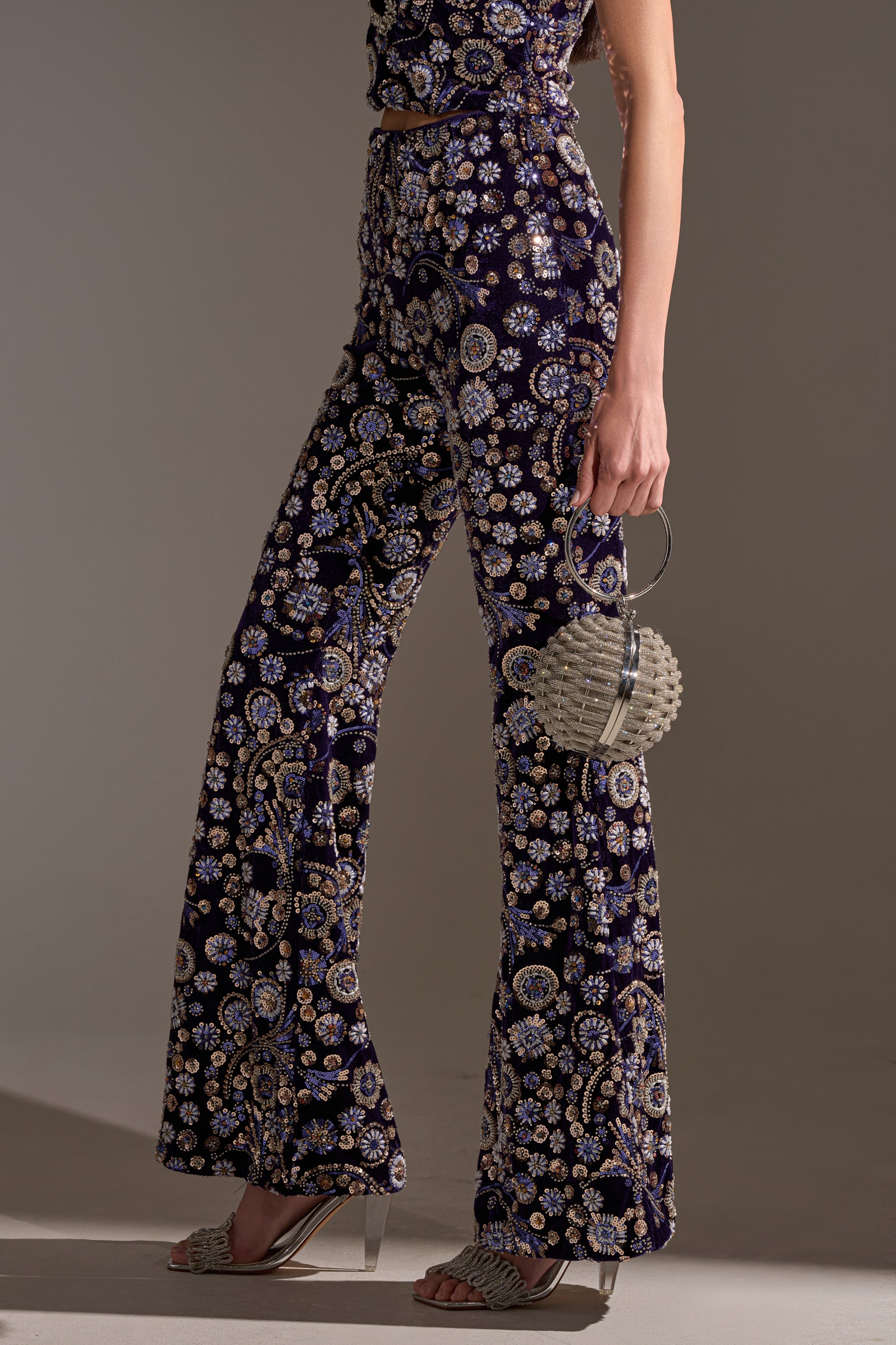A person stands in profile wearing the QUEEN OF EVERYTHING RHINESTONE TROUSER, featuring dark velvet fabric with a floral pattern. They hold a small round beaded handbag and wear fringe-detailed sandals with clear block heels.