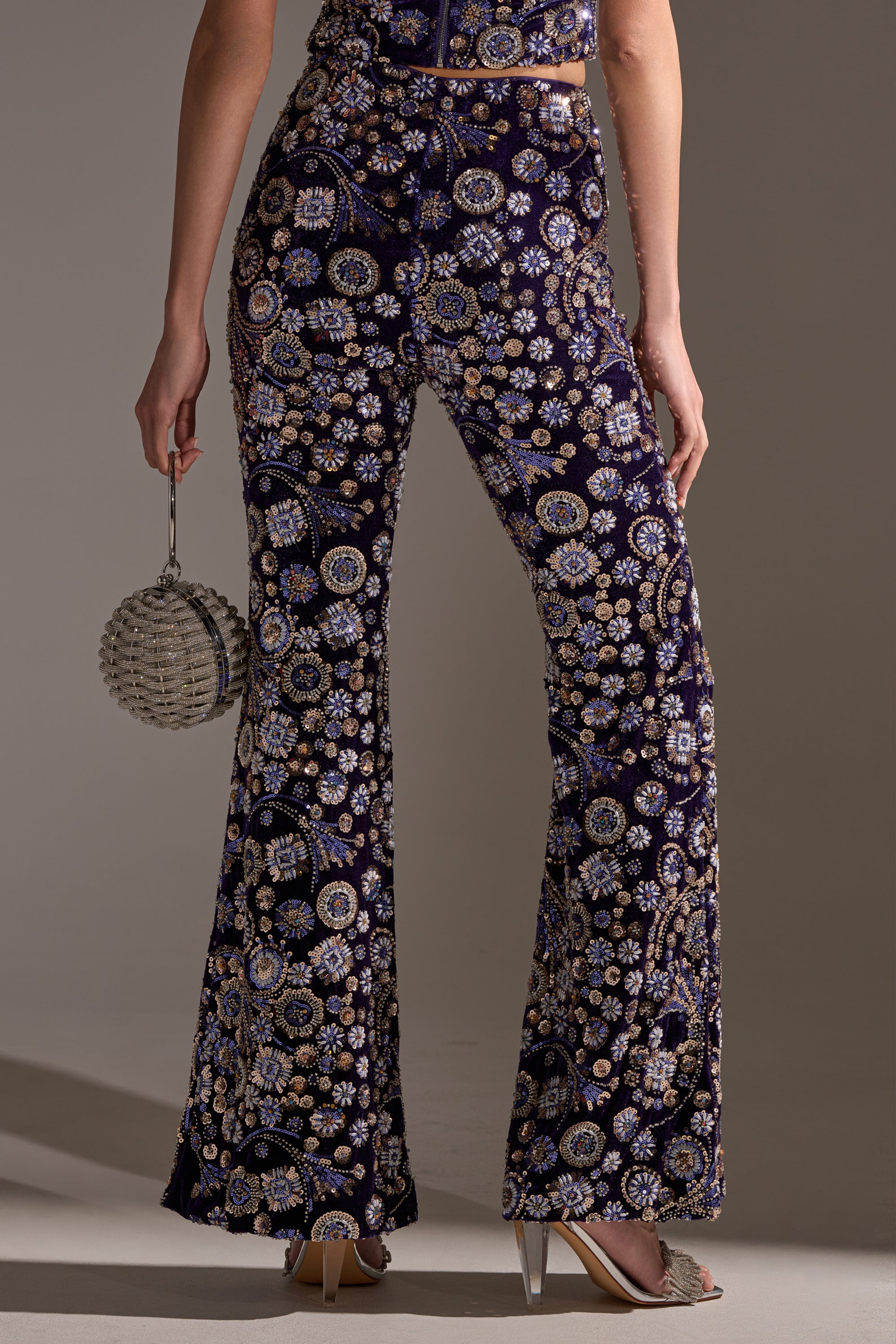 Wearing the QUEEN OF EVERYTHING RHINESTONE TROUSER—dark purple velvet pants with gold and silver floral embroidery and sequin accents—paired with a beaded round handbag and pointed high heels.